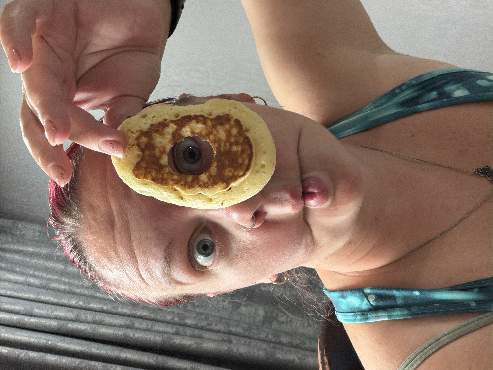 A woman is holding a pancake with a hole in the middle in front of her eye. The pancake is golden brown, and her eye is visible through the hole. She is wearing a blue and green tank top. Her nails are painted with glitter. The background is blurry.