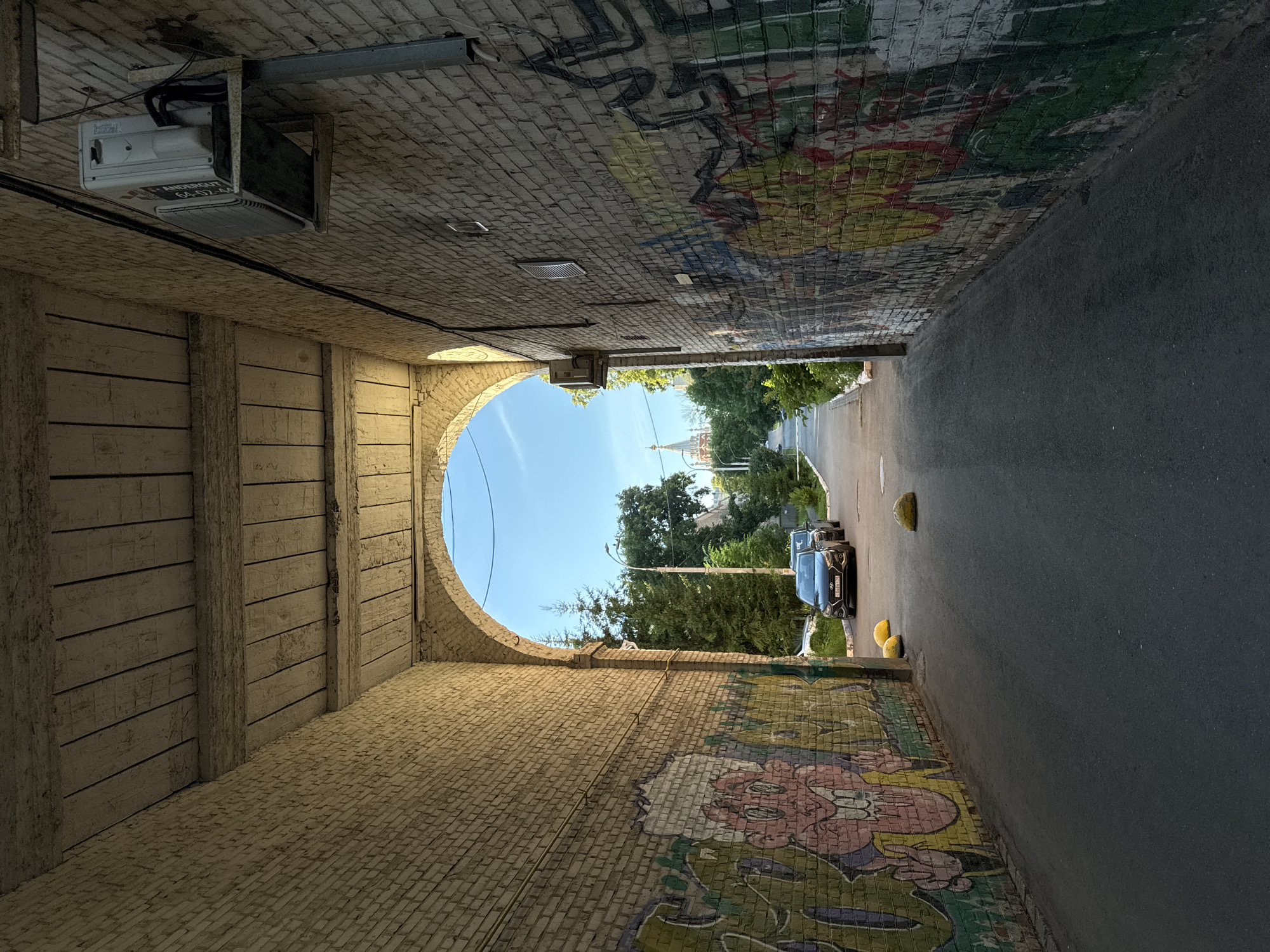 The image depicts a street tunnel in Izhevsk, Russia, viewed from within. The tunnel walls are made of brick and covered in graffiti. On the left side of the tunnel, there is a wooden structure. A light fixture is mounted on the ceiling of the tunnel. The tunnel opens up to a street with trees and a building with a dome in the distance. There is a car parked on the right side of the street, along with two yellow bollards. The sky is blue with a few clouds. The time of day appears to be daytime, possibly late afternoon, given the lighting. The overall mood is urban and slightly gritty due to the graffiti, but also has a sense of openness with the view of the sky and trees at the end of the tunnel. The graffiti on the walls includes various colorful drawings and lettering, but no specific readable text is clearly visible.