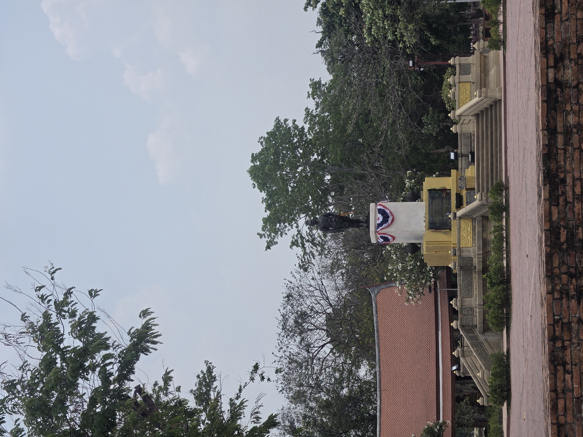 The image shows a statue in Phra Nakhon Si Ayutthaya City Municipality, Thailand, positioned on a pedestal, partially obscured by trees. The pedestal is adorned with gold accents and decorative elements. In the foreground, a red-tiled roof is visible, while a brick wall lines the right edge of the frame. The sky above is light blue, partially covered by clouds.