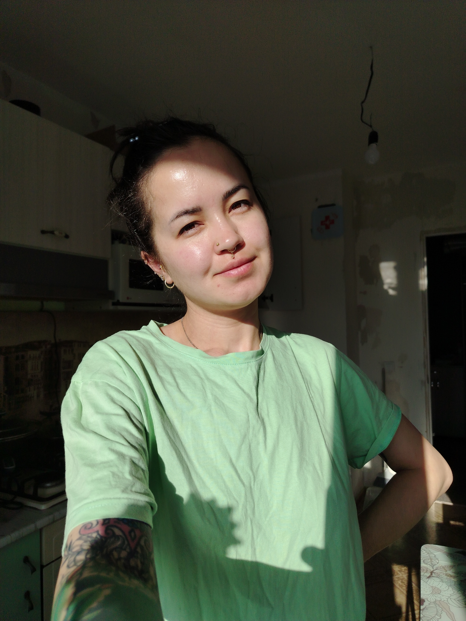 A person is in a kitchen. The person has dark hair pulled back, gold earrings, and a nose piercing. They are wearing a light green t-shirt. The background is a kitchen with cabinets, a microwave, and peeling wallpaper. A bare lightbulb hangs from the ceiling. Part of a tattoo is visible on the person's arm. The location appears to be a domestic interior. There is no evidence to confirm the location is in the city of Noviy, Russia.