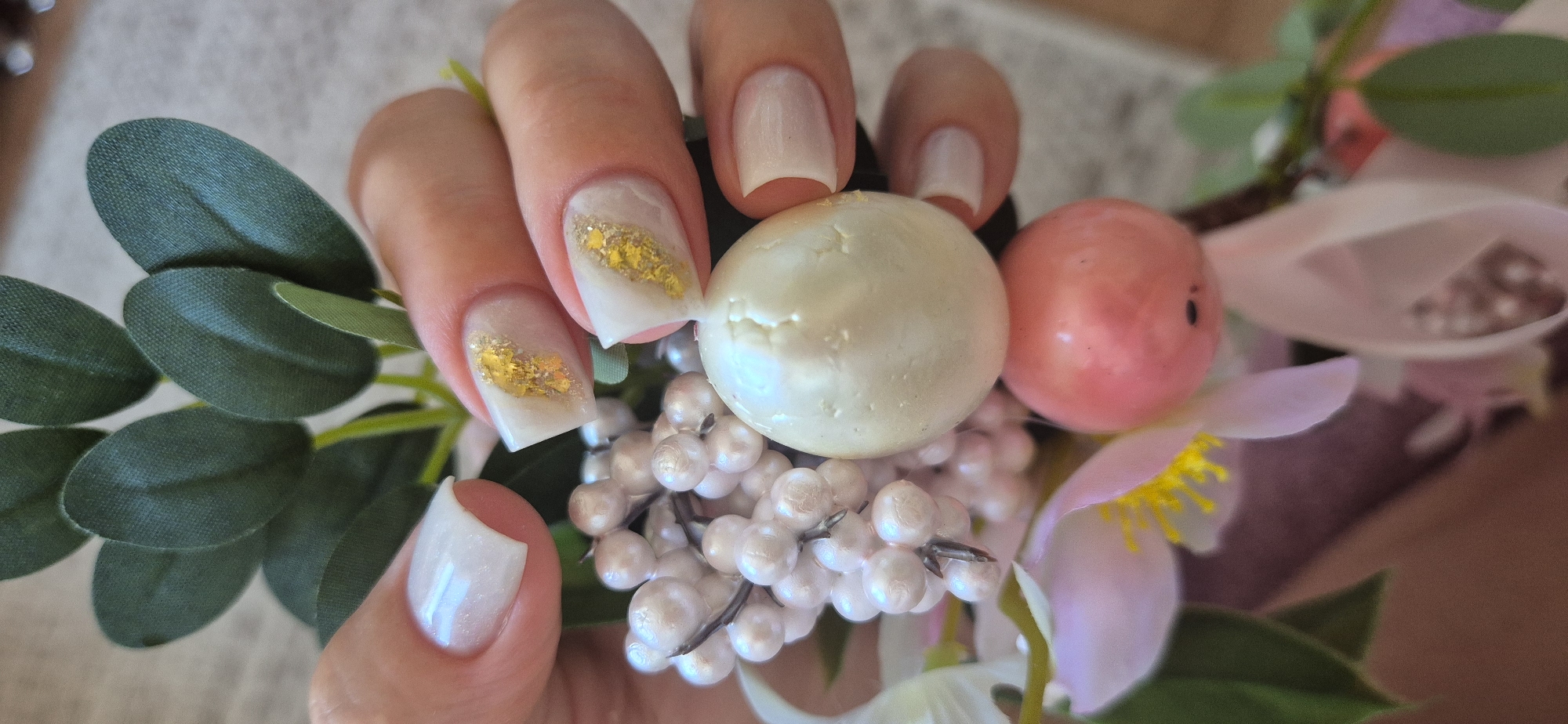 A close-up shot shows a hand holding a decorative arrangement. The hand has manicured nails, some adorned with gold foil accents over a white base. The arrangement includes green leaves, white and pink faux flowers, and various sizes of faux pearls. A large, pearl-like sphere rests against the fingers. The location is Unknown, Unknown.