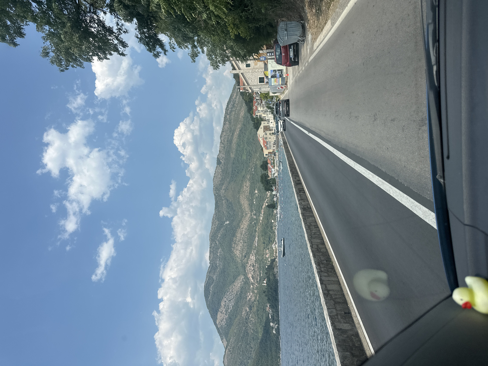 The image shows a scenic view of a coastal road in Donja Lastva, Montenegro, during the daytime. The perspective is from inside a vehicle, looking forward through the windshield. A rubber duck toy is visible on the dashboard in the foreground.
The road stretches ahead, curving slightly to the right, with a white line marking the lane division. To the left of the road is a stone retaining wall bordering a body of water, likely the Adriatic Sea. Several boats, including a small speed boat, are anchored or moving on the water.
In the background, a steep, verdant mountain range dominates the landscape. Along the coast, nestled at the foot of the mountains, are buildings with red-tiled roofs, suggesting a small town or village. The sky is a clear blue with scattered white cumulus clouds.
On the right side of the road, there are trees and foliage. Several cars are driving on the road, and some parked vehicles are visible near the buildings. Two large, grey refuse bins are placed on the side of the road near a building. The overall impression is a sunny day in a picturesque coastal area.
