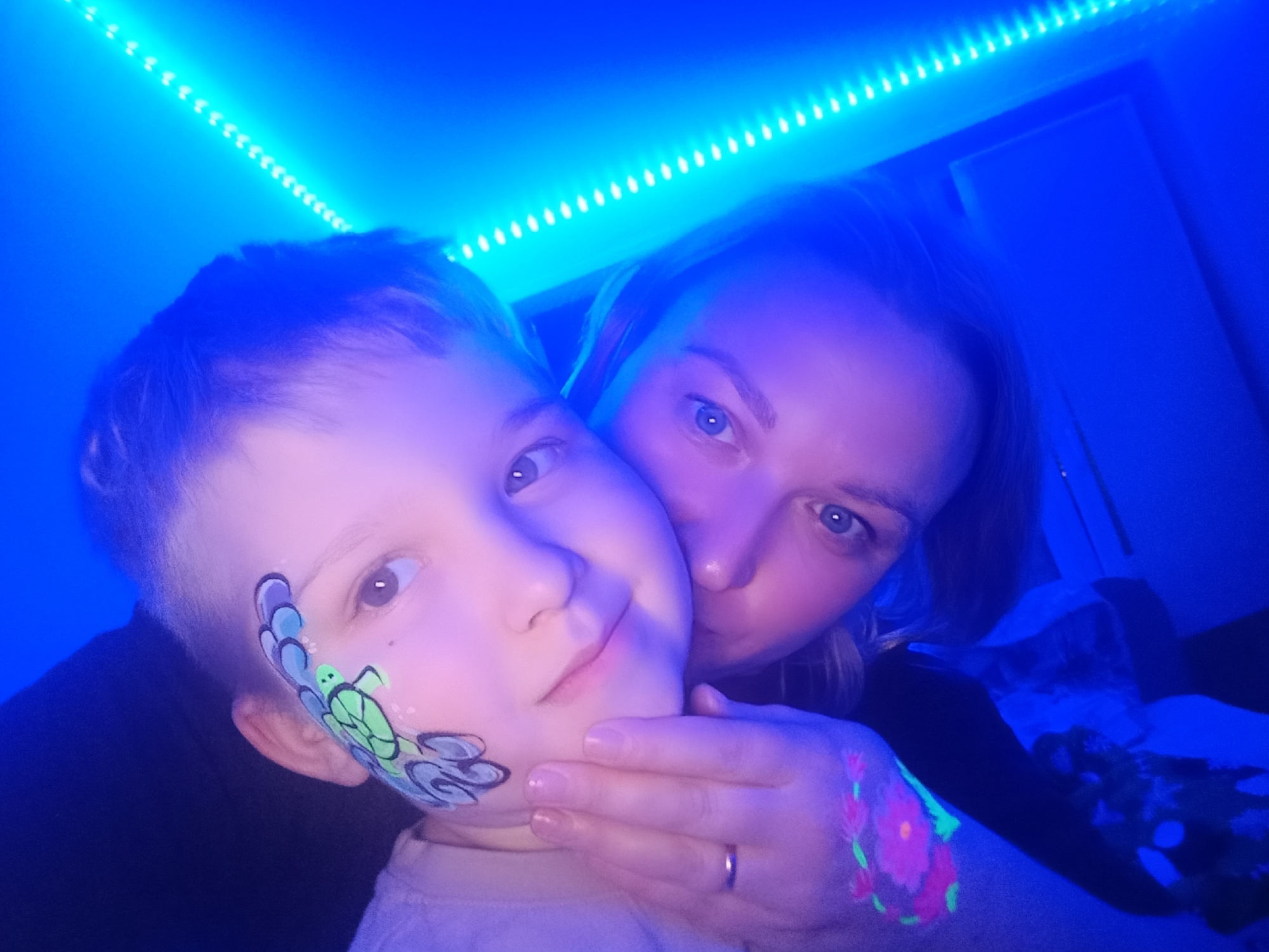 This image captures an intimate moment between an adult woman and a young child in Chaikovsky, Russia, illuminated by vibrant blue LED strip lights. The scene appears to be set indoors during nighttime or in a specially darkened room, suggesting a celebratory event or party atmosphere.
The young child, positioned prominently in the foreground, features artistic face paint on their left cheek, depicting a green turtle surrounded by blue and purple wave-like designs. The child looks towards the camera with a soft, gentle expression. The woman, likely the child's mother, gently holds the child's face, her hand resting on their cheek. She has a tender, affectionate gaze directed at the camera, and a simple gold band is visible on her left ring finger.
A striking detail is the glowing body paint on the woman's right arm/hand, showcasing a bright pink and green floral design that fluoresces under the blue lighting. This, along with the child's face paint, indicates a glow-in-the-dark or UV-reactive theme for the occasion. Bright blue LED strip lights are visible along the top of the frame, casting a cool, energetic glow that defines the overall mood and highlights the subjects. The general feeling conveyed by the image is one of warmth, affection, and celebration. No text is visible in the image.