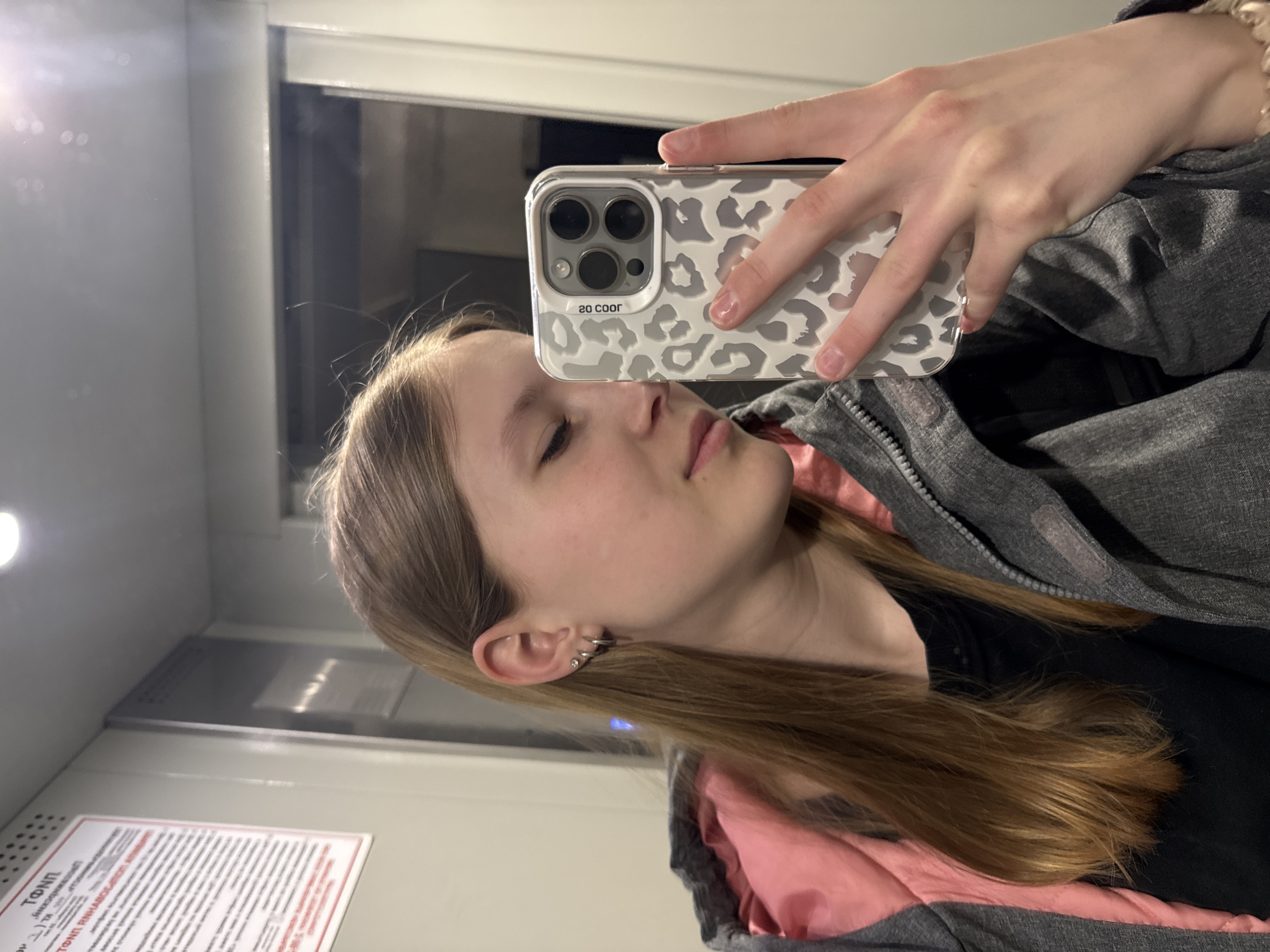 A young woman is taking a selfie with a smartphone that has a white case with a gray leopard print pattern. She is indoors, likely in a train car, as suggested by the window, interior paneling, and sign with Russian text. She has light brown hair, fair skin, and is wearing multiple earrings. She is wearing a dark shirt and a gray jacket with pink lining. The location is presumed to be in Russia, given the language on the sign. The city of Chaikovsky cannot be definitively determined from this image, but the sign may provide further clues for that assessment.