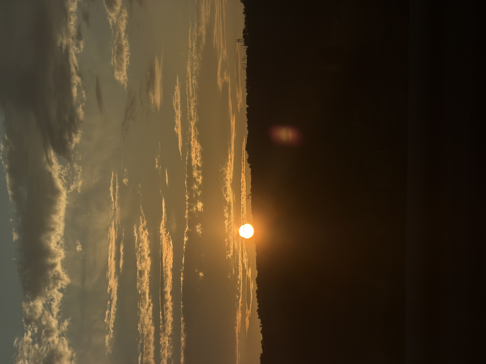 The image captures a sunset over a silhouetted landscape. The sun, a bright orb, is positioned in the upper left quadrant of the frame, casting a warm, golden glow across a sky filled with layered clouds. The clouds vary in color from pale yellow to dark gray, with some illuminated by the setting sun and others casting shadows. Below the sky is a dark, undulating silhouette of a treeline, suggesting rolling hills. In the foreground, the scene is dominated by a dark, undefined expanse, possibly a body of water or a shadowed plain, with a faint horizontal line suggesting the meeting of land and sky. A lens flare, appearing as a reddish-orange circular smudge, is visible in the lower right portion of the image, obscuring a small part of the treeline. There are no visible people or specific man-made structures in the immediate foreground or midground, although very faint outlines of what might be distant power line towers are discernible against the sky along the crest of the treeline. The overall impression is one of natural tranquility and the dramatic beauty of a sunset.