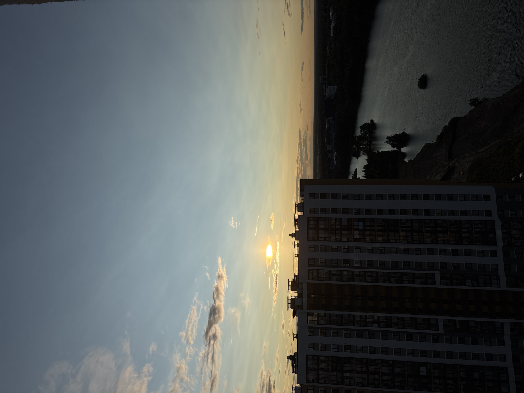 An aerial, high-angle, wide shot captures a serene cityscape at sunset. The sun, a brilliant orb of light, hangs low in the sky, casting a warm glow across the horizon. Fluffy clouds are scattered across the expanse of the sky, tinged with hues of orange and pink. Dominating the left foreground is a tall, multi-story building, its facade a mosaic of windows and geometric patterns. To the right, a tranquil body of water stretches out, its surface reflecting the soft light of the setting sun. Islands dot the water, adorned with clusters of trees. The distant cityscape is shrouded in a veil of shadow, with faint outlines of buildings and lush greenery discernible against the darkening sky.
