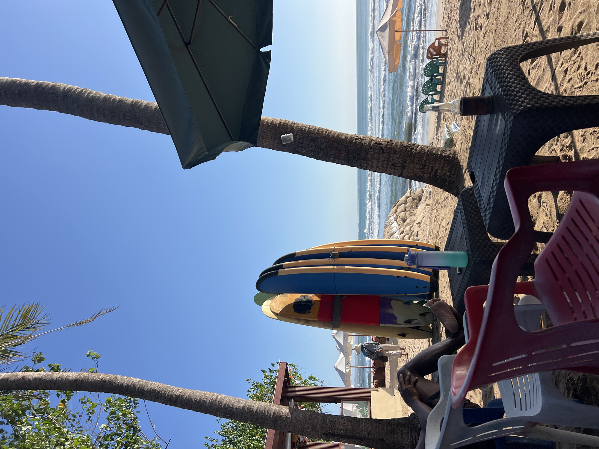 In Kuta, Indonesia, a beach scene unfolds under a clear blue sky. In the foreground, plastic chairs in red and white are set on the sand. A person's legs and feet are visible, resting on one of the white chairs. Adjacent to the chairs, a dark plastic table holds a bottle of brown liquid and a blue and green water bottle. Further back, several surfboards are propped up on the sand, displaying a variety of colors and designs, including yellow, red, blue, and stripes. To the left, a wooden structure with a brown roof and white walls stands partially in view, with palm trees providing shade. A person wearing a light-colored hat and matching pants and jacket walks away from the surfboards toward the beach. The ocean waves gently roll onto the sandy shore, and several beach umbrellas and chairs are scattered along the coastline. Another palm tree stands prominently on the right side of the image, with a dark green umbrella extending from it. The scene evokes a relaxed atmosphere on a sunny day at the beach.