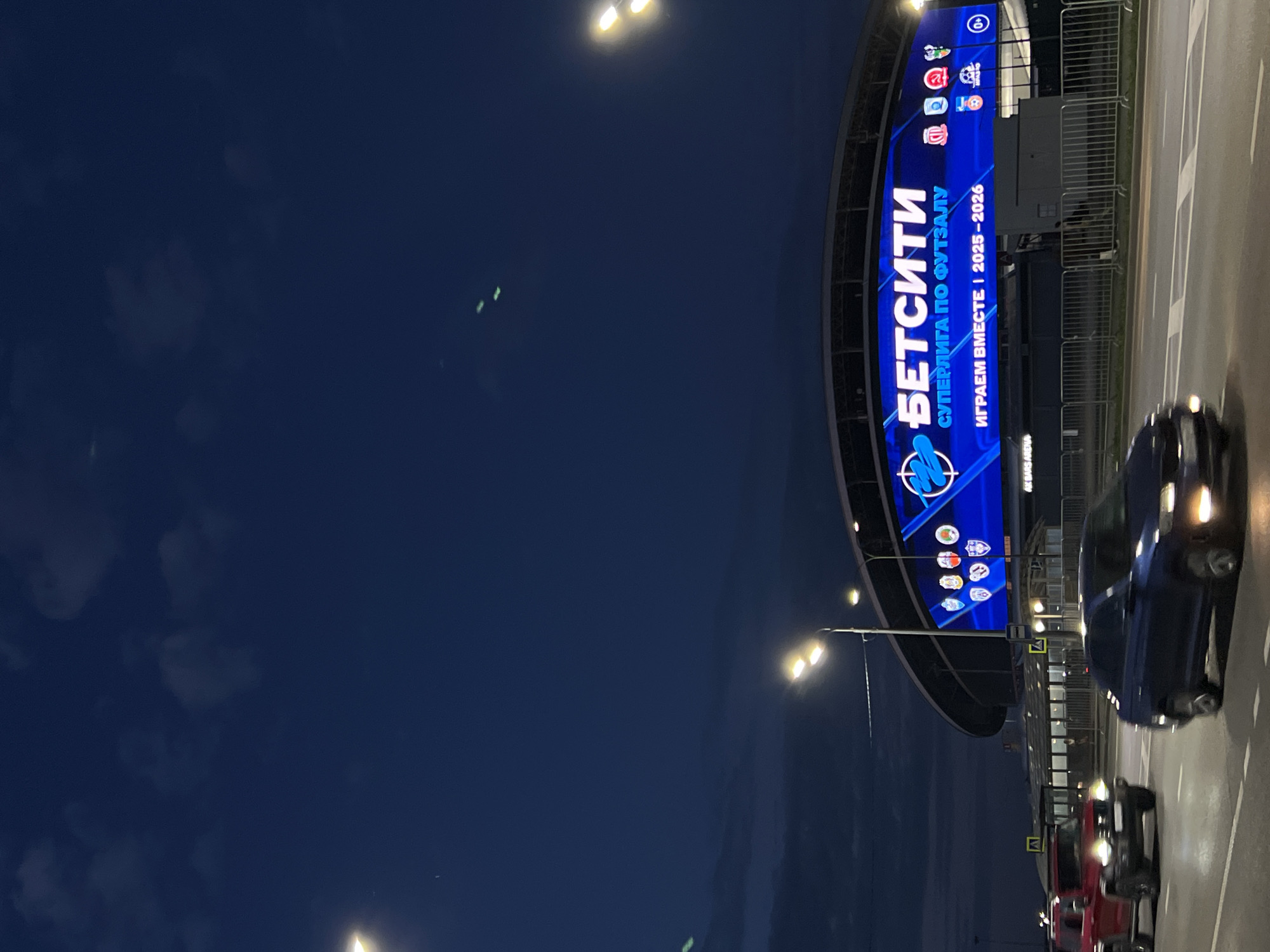 The image shows a large LED screen on a stadium that says "БЕТСИТИ" (BETCITY) and "СУПЕРЛИГА ПО ФУТЗАЛУ" (FUTSAL SUPER LEAGUE). Below that, it reads "ИГРАЕМ ВМЕСТЕ | 2025-2026" (WE PLAY TOGETHER | 2025-2026). The screen also displays various team logos. In front of the stadium, there are two cars driving on a road at night. The sky is dark blue with some clouds and streetlights are visible.