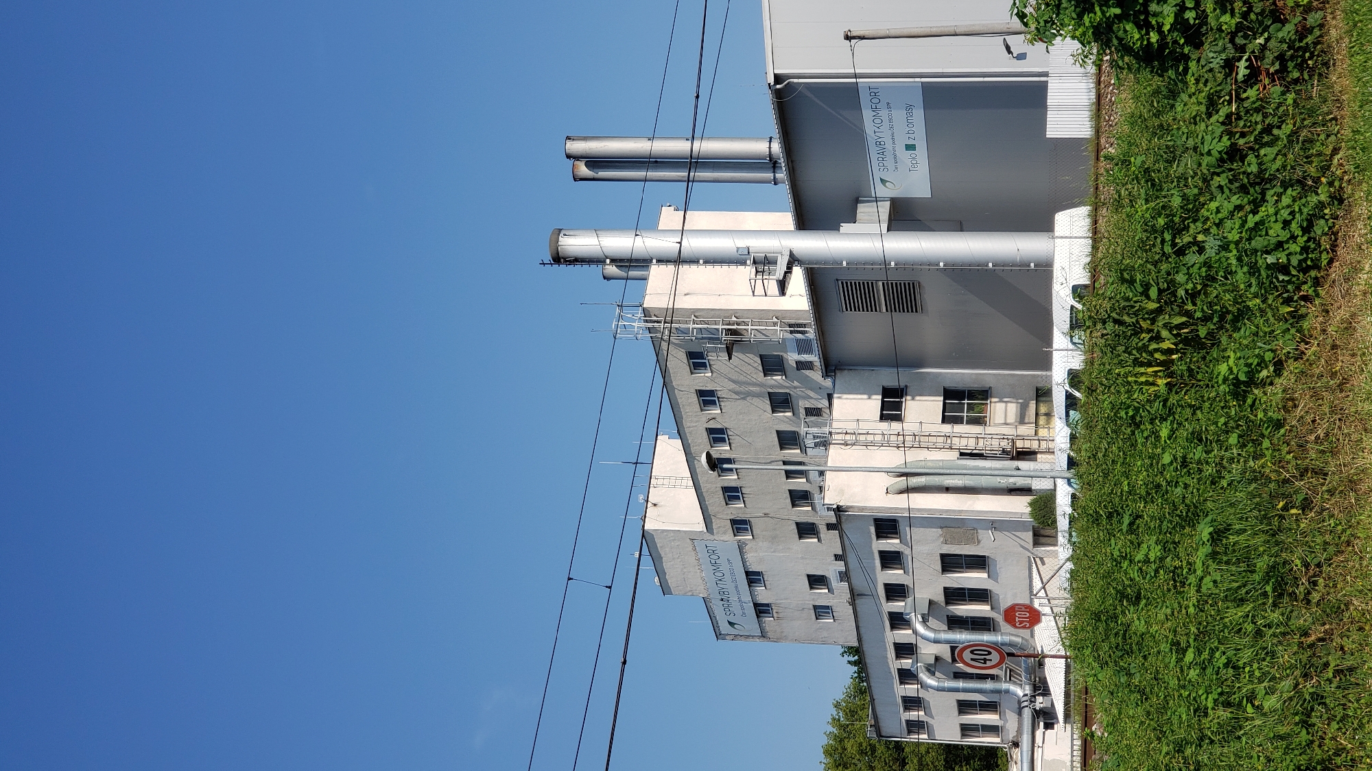 The image shows a factory building with multiple levels and a tall smokestack. The building is white with many windows. There are signs on the building that read "SPRAYBYTKOMFORT" and "Teplo z bomasy". There are also traffic signs visible, one indicating a speed limit of 40 and another a stop sign. The building is located next to train tracks and is surrounded by lush green vegetation in the foreground. The sky is clear and blue. The factory is the headquarters of SPRAYBYTKOMFORT s.r.o., a company that provides energy services, including heating and cooling. The company is located at Kuzmányho 8, Prešov, Slovakia.