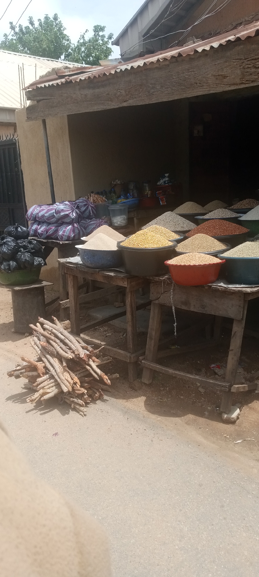 The image depicts an outdoor market scene likely located in Jos, Nigeria. The focal point is a stall selling grains and pulses, with large basins filled with different types of grains arranged on wooden tables. There are at least seven basins visible, containing grains of various colors including beige, yellow, red, and green, suggesting a variety of offerings. To the left of the table are bundles of firewood resting on the ground, and additional bags of goods are piled on a table and in a green basin. The stall is covered by a simple corrugated metal roof. The street area in front of the stall is a light-colored asphalt.