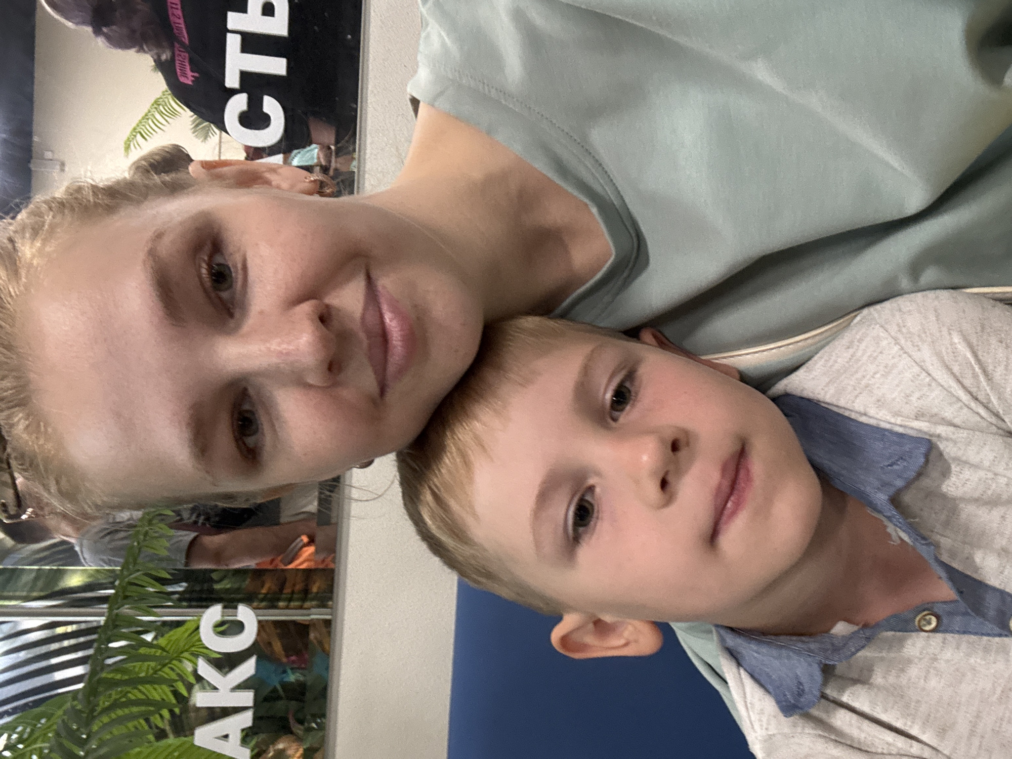 An outdoor, medium shot captures a woman and a young boy in what appears to be a selfie. The woman is on the right, with her blonde hair pulled back, wearing a light green t-shirt. The boy, on the left, has blonde hair and is wearing a light grey shirt with a blue collar. He is leaning against the woman. In the background, a large sign with white letters, partially visible, reads "АКС". Reflections in the background show other people and possibly a store interior with plants. The overall lighting suggests an indoor setting with possibly natural light entering from the background.