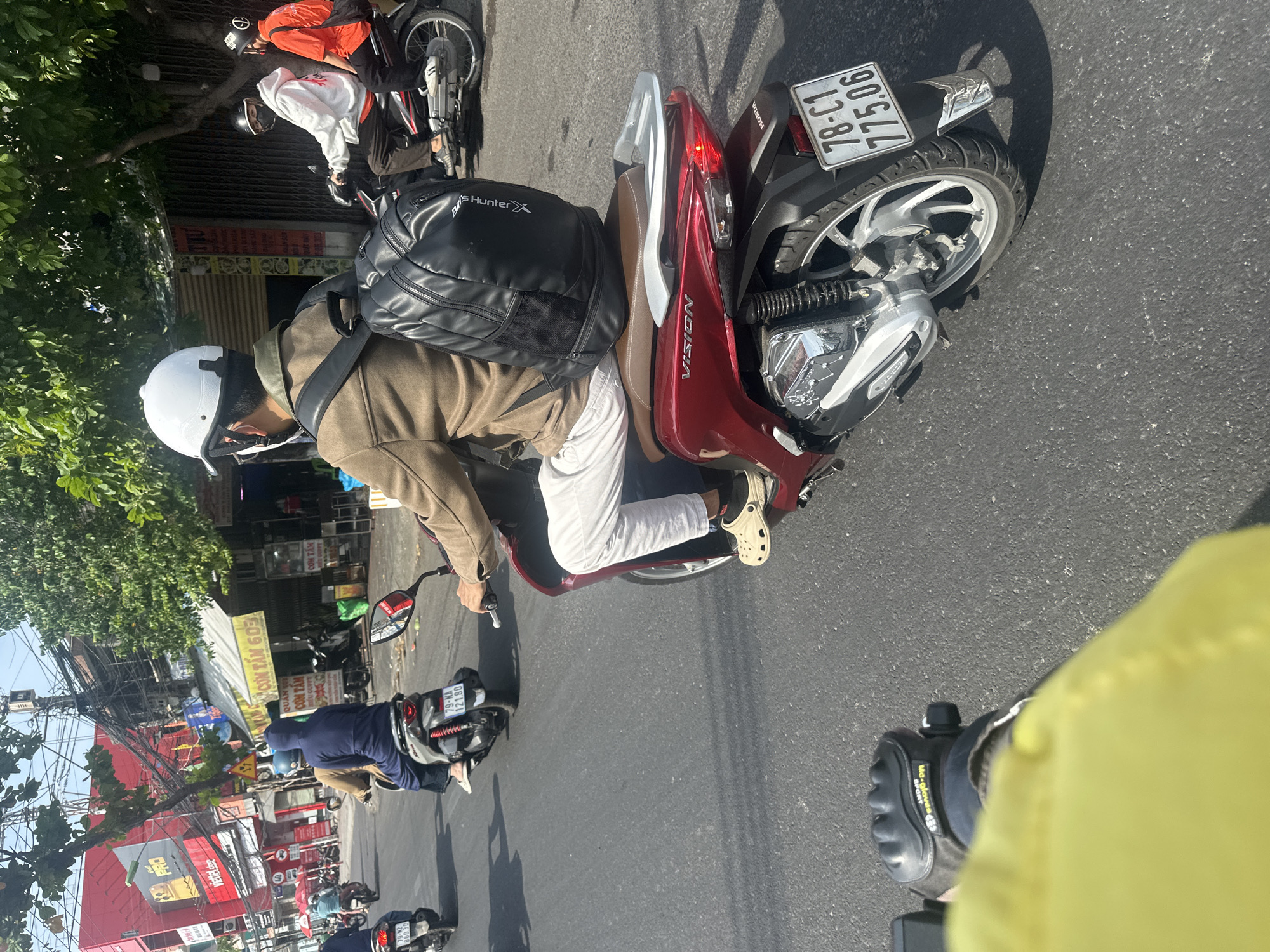 In Bac Nha Trang Ward, Vietnam, on a sunlit day, a street scene unfolds with multiple motorcycles navigating a roadway. The foreground shows the leg and arm of someone wearing a yellow jacket and black motorcycle gloves, as well as the rear of a red Honda Vision scooter with a license plate reading "78-C1 775.06". A person on this scooter wears white pants and tan Crocs. To their left, another scooter carries a person in a blue hooded jacket. Further ahead, a person in a brown jacket with a black backpack is riding a scooter. Other motorcycles and riders are visible in the background, including one wearing a white helmet and another in an orange shirt. The environment includes trees, buildings with signage such as "COM TAM 603", and utility poles with many wires. The asphalt road shows some debris.