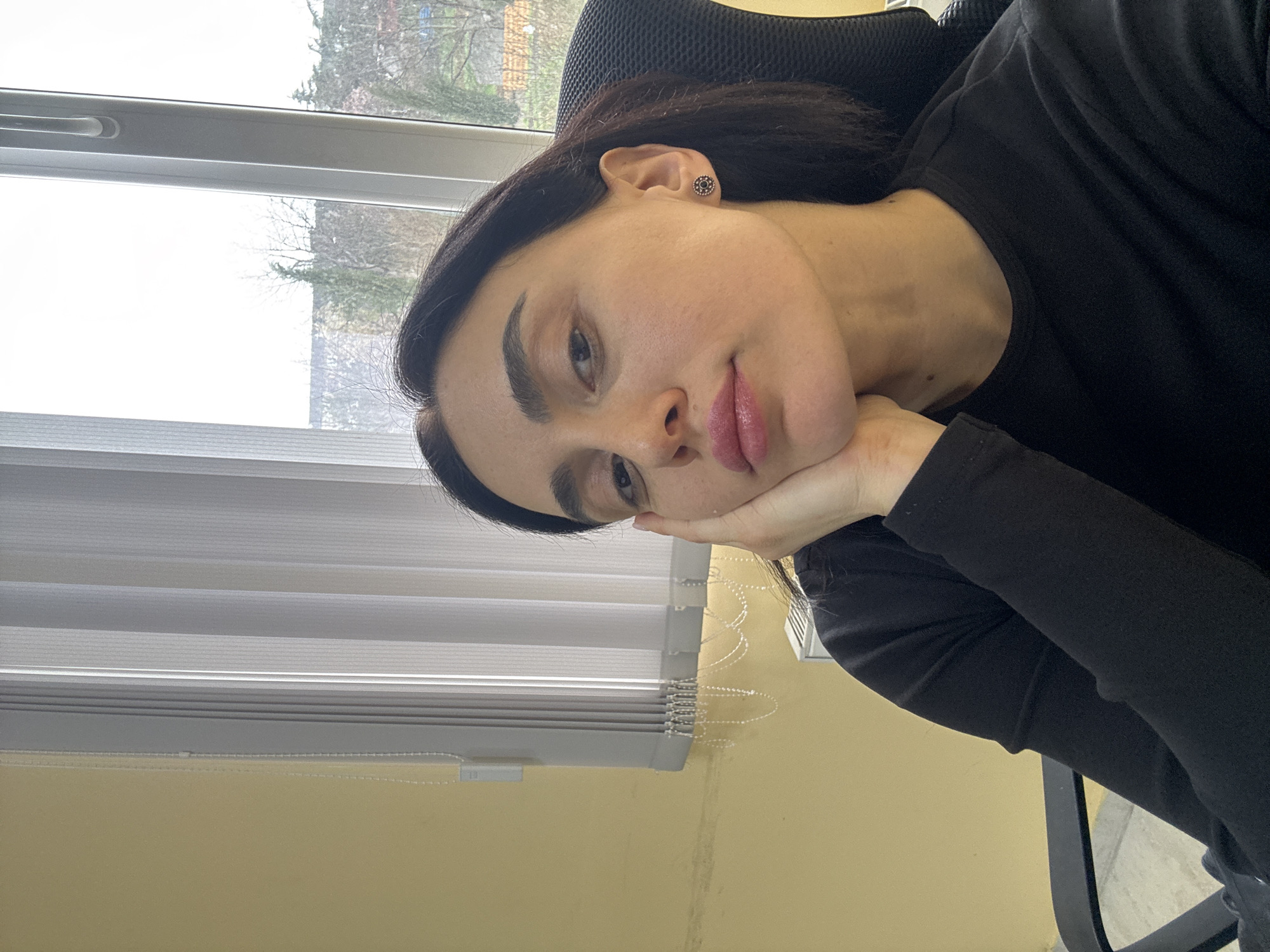 A young woman with dark hair and defined eyebrows is resting her head on her hand. She is wearing a black long-sleeved shirt and a small earring. The background includes a window with partially closed blinds, revealing trees outside. She appears to be sitting in an office chair. The image was taken in a location that could be the city of Tbilisi, Georgia, given the user's instruction.