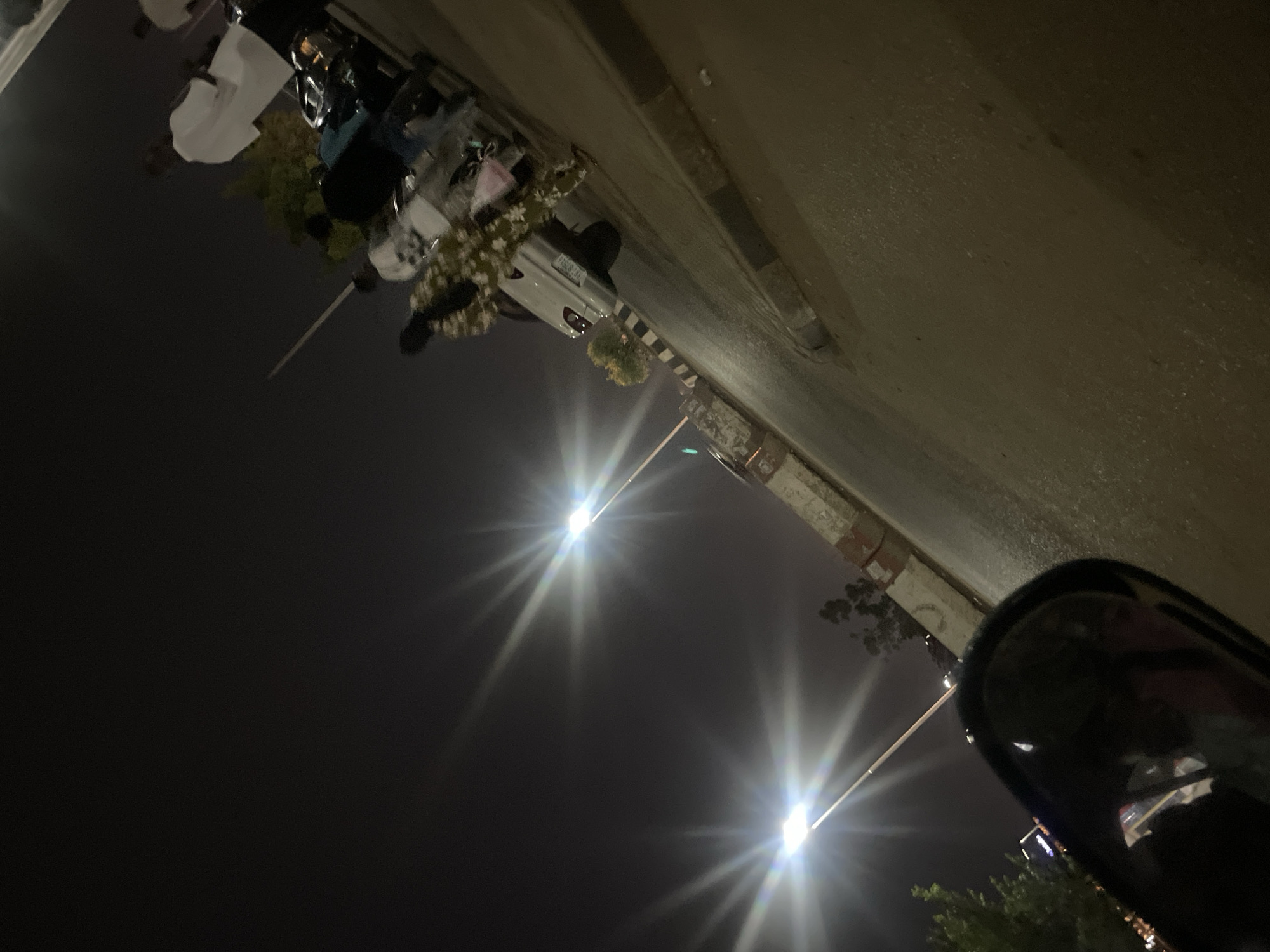 The image captures a nighttime street scene, rotated 90 degrees counter-clockwise. When viewed upright, a dark asphalt road extends into the distance on the right, alongside a light-colored concrete or stone curb and pavement on the left.
Two prominent, brightly lit streetlights with strong starburst effects are visible, illuminating the dark sky. One streetlight is in the mid-ground, and another is further down the street. A white car is parked on the left side of the road, partially on the pavement. Several people are gathered near the car; their details are obscured by the low light, but one person appears to be wearing a patterned garment, possibly green and white. Scattered foliage, including bushes or trees, is visible along the pavement and near the car.
In the extreme foreground on the bottom right (when upright), a blurred part of what appears to be a side-view mirror of a vehicle can be seen. The license plate on the white car is visible and reads "DV-88931". The scene is quiet, typical of an urban or suburban street at night. No specific weather conditions or emotions are discernible.