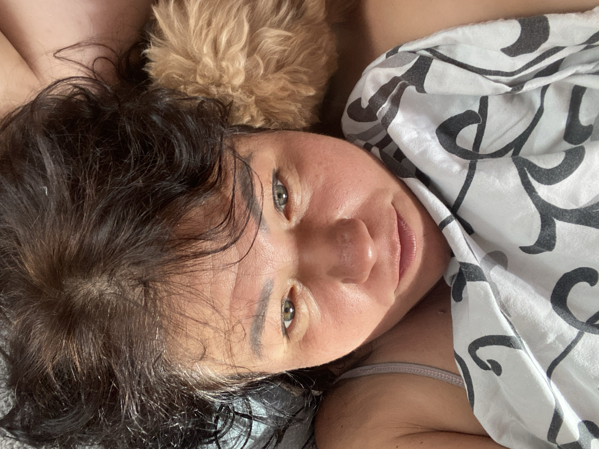 A woman with dark, wavy hair is lying down, possibly in bed, with a light-colored blanket with a black and gray abstract pattern draped over her chest and shoulder. Part of her face is visible, with her eyes looking towards the camera. She appears to be indoors, and her surroundings are not clearly identifiable. A small section of what appears to be a tan-colored dog's fur is visible near her head. The image was captured in Dnipro, Ukraine.