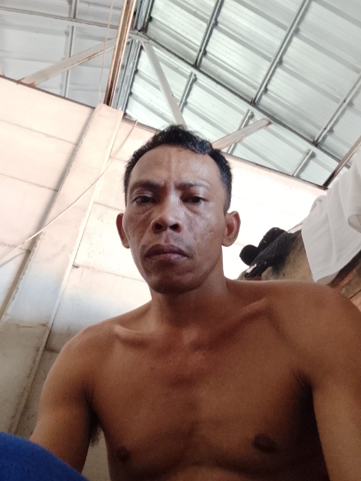 A shirtless man is seen from the chest up, in an interior space that appears to be a simple structure with a metal roof and white walls. The location is potentially in Bukit Ketok, Indonesia. The man has dark hair and skin, and a neutral expression. Clothes hanging in the background.