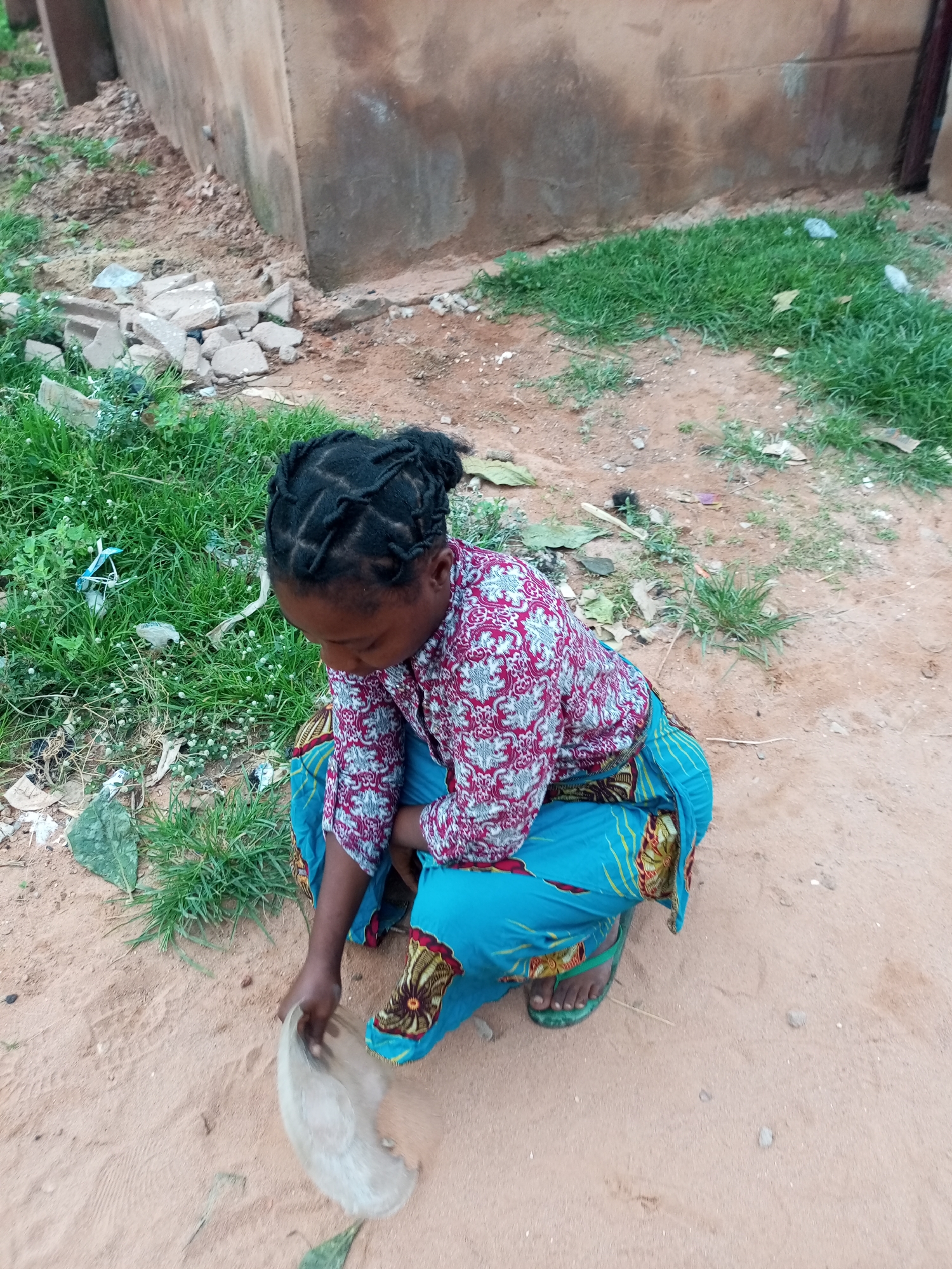 A young Black female, appearing to be of Nigerian descent, is captured in a high-angle shot, crouching on sandy ground next to a building. She is wearing a long, blue wrap skirt adorned with a colorful, patterned fabric and a long-sleeved, red and white patterned shirt. Her dark hair is braided and styled in a bun on top of her head. Her right hand is extended, holding a piece of translucent plastic. The environment appears to be an outdoor urban setting in Tempure, Nigeria, with patches of green grass and scattered debris visible around the building's foundation. The building itself is made of a light brown, textured material, suggesting it might be an older structure. The ground is a mixture of sand and dirt, with some small rocks and dried leaves present. The overall scene depicts a moment of environmental tidiness as the girl collects discarded plastic.