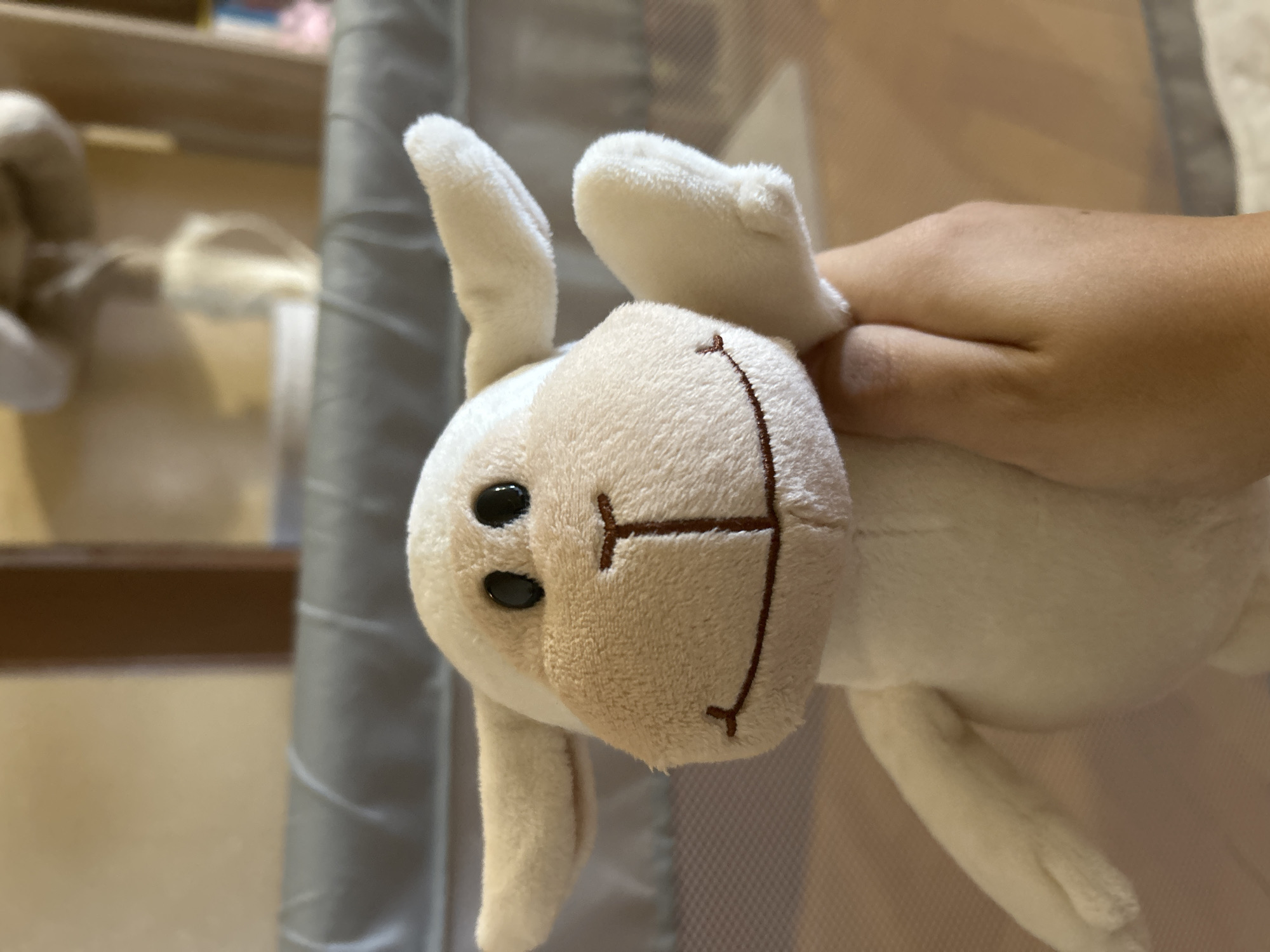 A human hand with light-to-medium skin tone holds a white plush stuffed animal, which resembles a lamb or sheep, in an indoor setting. The stuffed animal has black, oval-shaped eyes, a tan-colored muzzle with a stitched brown inverted 'T' for a nose and a curved line for a smile, and two floppy ears. Its left arm is raised, suggesting a waving gesture. The hand, visible in the lower right, grips the toy's torso. In the immediate background, a grey, padded or fabric-covered railing or edge is present, possibly belonging to a playpen or crib. The background beyond this railing is significantly blurred, showing indistinct light-colored furniture or wall elements and a faint, light-colored object suspended in the upper center, possibly part of a baby mobile or hanging toy. The specific location in the city of Sofia, Bulgaria, cannot be determined from the visual information provided in the image.