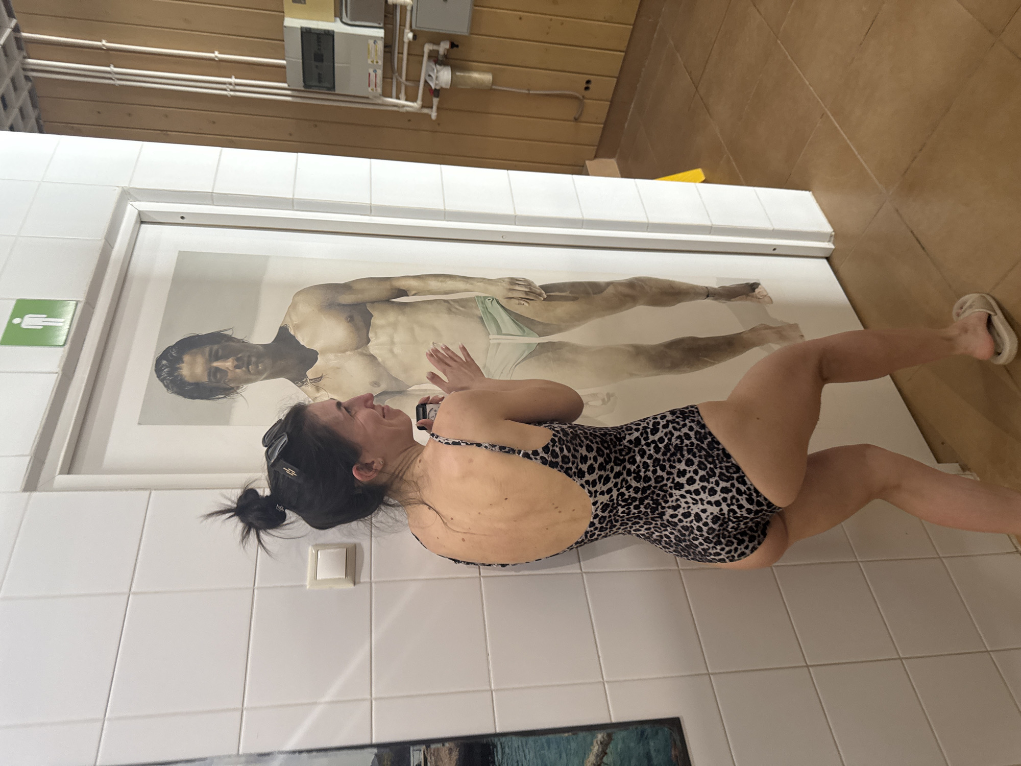 An image captures a woman in an indoor setting, likely a spa or changing room, interacting with a large framed artwork. The woman, with dark hair tied back and sunglasses perched on her head, lies on her back on what appears to be a tiled or white surface. She wears a leopard-print one-piece swimsuit and light-colored flip-flops are visible on her feet. Her left hand rests on her chest, while her right hand holds a cell phone near her ear or chest. She is looking upwards towards a large, framed print or painting depicting a shirtless man, possibly partially submerged in water, wearing light-colored swim briefs. The man in the artwork has dark hair and a tattoo on his left arm. The woman’s left hand appears to playfully reach towards the man's midsection within the artwork, creating an illusion of interaction.
The background features white tiled walls on the lower section, transitioning to wooden paneling higher up. A green sign with a white stick figure, commonly indicating a restroom or changing room, is affixed to the white tiles. Exposed pipes and various electrical boxes are visible on the wooden wall. The floor is covered in light brown or beige square tiles. The overall ambiance suggests a functional, possibly older, public facility. The woman appears relaxed, possibly amused or contemplating the artwork. No other discernible text is visible besides the restroom sign.