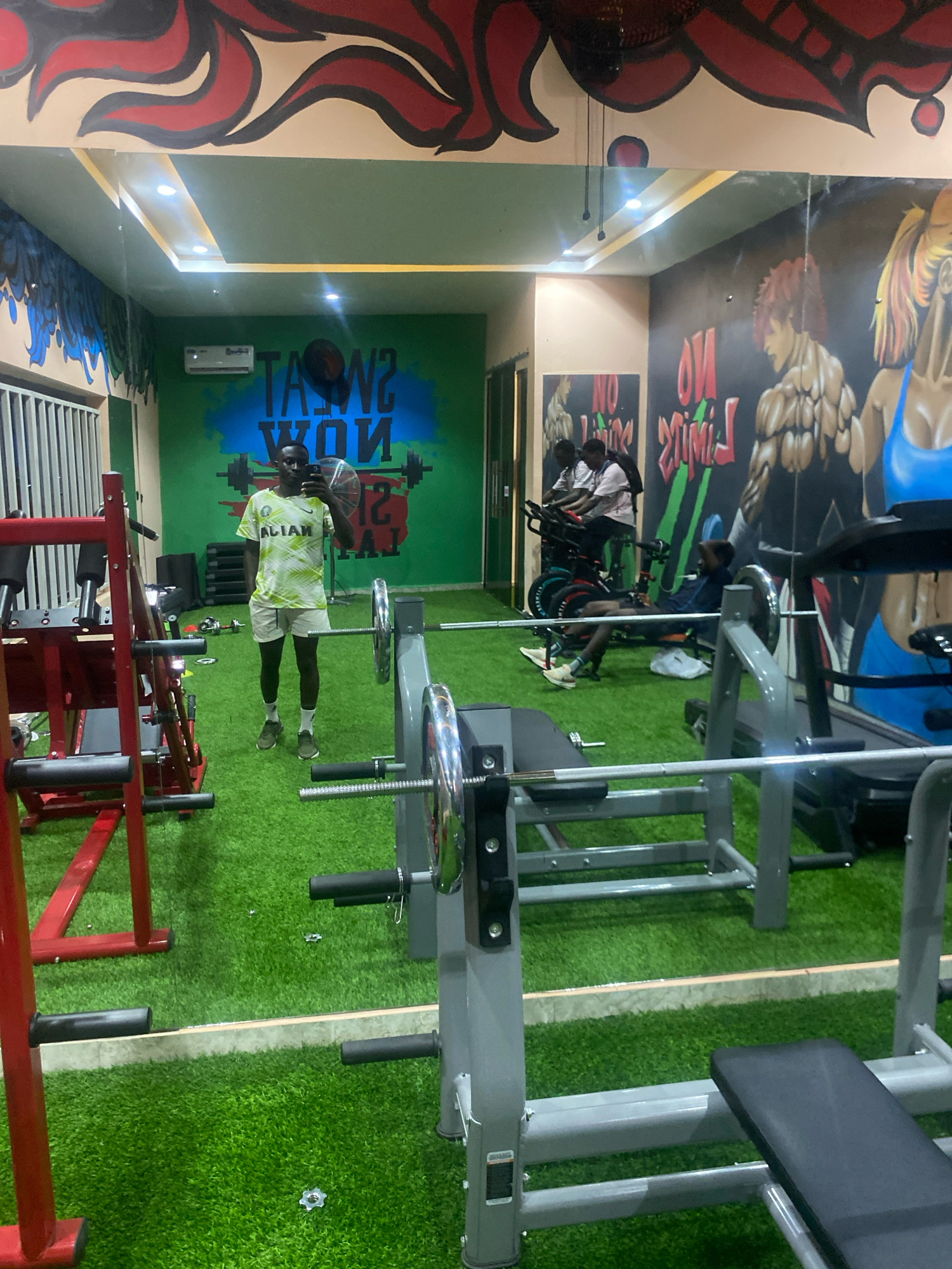 This image depicts the interior of a gym in Barden Yana, Nigeria. The gym features artificial turf flooring and a mural on the wall with the text "SWEAT NOW." Several pieces of gym equipment are visible, including weight benches and exercise bikes. In the foreground, a person is taking a mirror selfie, wearing a light-colored t-shirt and shorts. In the background, two individuals are using stationary bicycles, and another person is seated on a weight bench. The environment appears to be well-lit with overhead lights.