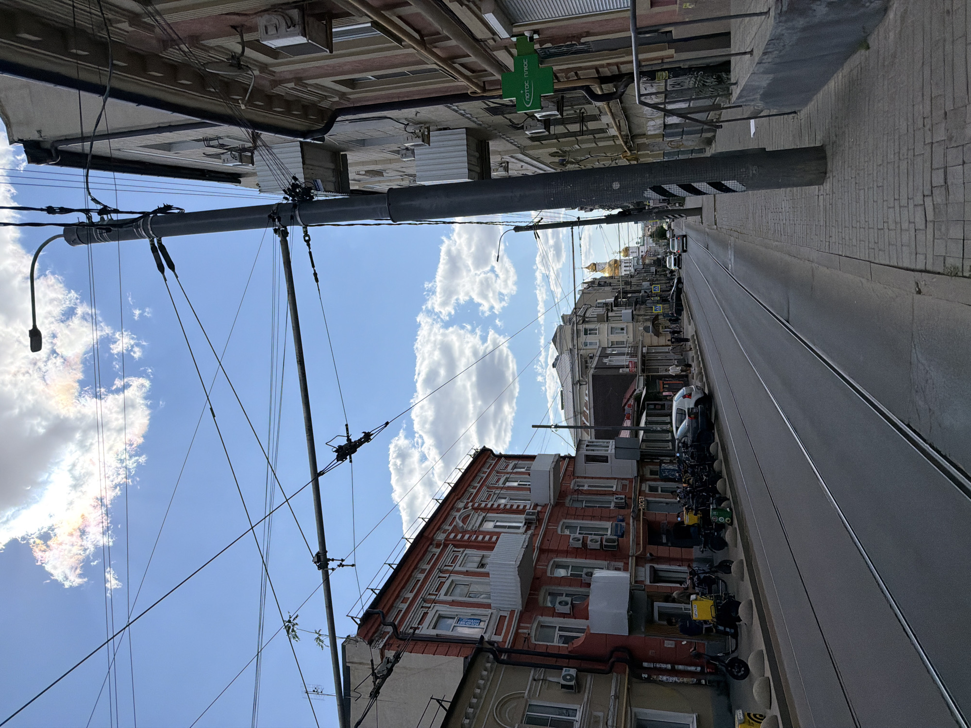 A street scene in Rostov-on-Don, Russia, features a tram line running down the center of the road. On the left, a red building with white trim and balconies is visible, with several electric scooters parked along the sidewalk. Further down the street, a white car is parked on the right side. The right side of the street is lined with buildings, one of which has a green pharmacy sign with a white cross. Overhead, numerous electrical wires crisscross the sky, supported by tall poles. In the distance, a golden-domed church is visible. The sky is blue with scattered white clouds. Several people are walking on the sidewalks, and vehicles are present on the road.
