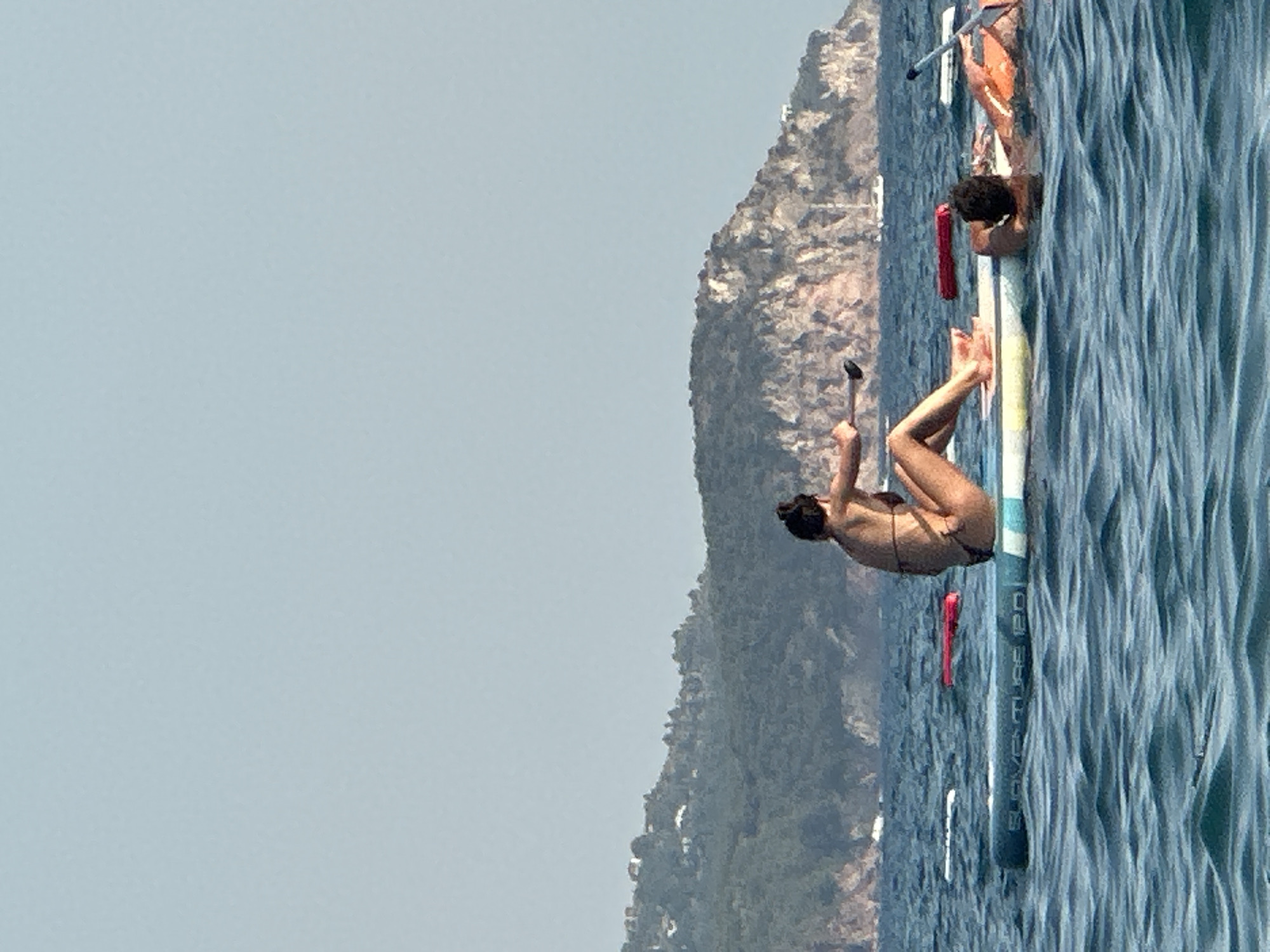 In the waters near Boreti, Montenegro, a woman is seated on a stand-up paddleboard, holding a paddle. She is wearing a dark bikini. To her right, another person is lying on a paddleboard, their head resting on their arms. They are wearing an orange life vest. The water is a deep blue with visible ripples. In the background, a green, hilly coastline with scattered buildings rises from the sea, under a clear, pale blue sky. Several buoys are visible in the water.