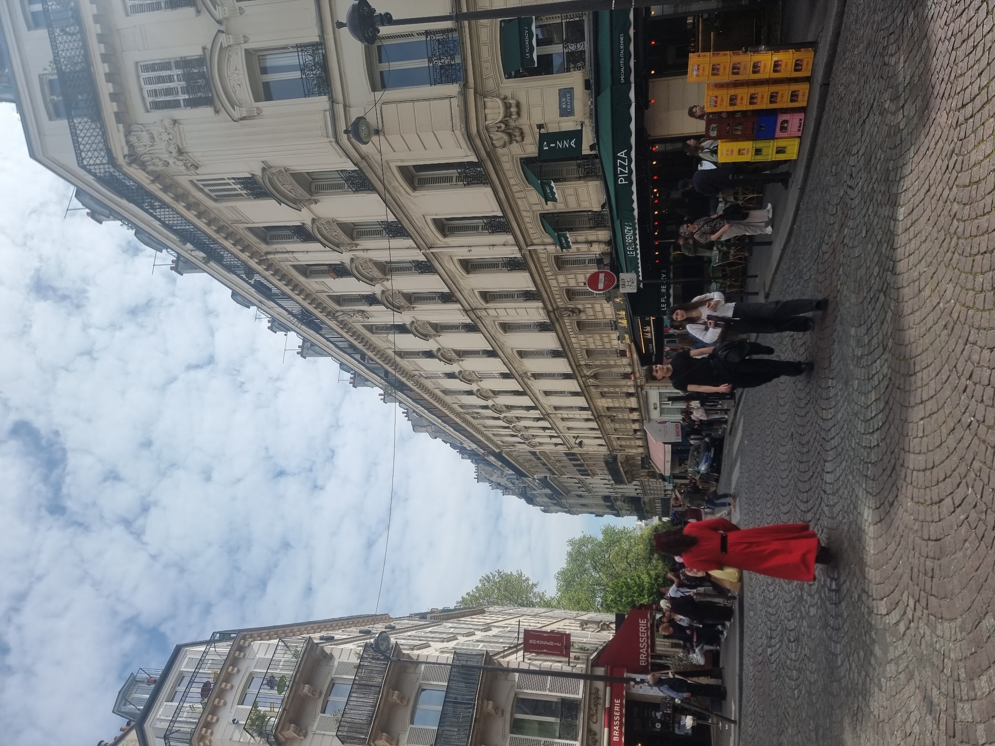 The image captures a vibrant street scene in Paris, France, featuring classic Haussmannian architecture and daily life under a cloudy sky. The perspective is tilted, showing tall, ornate buildings lining a cobblestone street.
In the foreground and midground, several people are visible. A woman in a striking red dress walks across the cobblestones, moving away from the viewer. Other individuals are seen strolling, standing, or seated at outdoor tables of restaurants.
The street is bustling with commercial activity. On the right, a multi-story building houses "LE FLORENZY," a restaurant identified by green awnings proclaiming "PIZZA" and "SPÉCIALITÉS ITALIENNES." Outside, stacks of colorful crates filled with "Coke" are neatly arranged. A "RUE CHAPPE" street sign is affixed to the building, indicating the specific location. Further down, another establishment has a sign for "BACHIR." On the left, another building features "BRASSERIE" signs, with one specifically mentioning "Café Chappe." A red traffic sign with "SAUF" and a bicycle symbol is also visible.
The architecture is notable for its intricate details, including decorative balconies with wrought-iron railings and numerous shuttered windows. The sky is overcast with fluffy white clouds, suggesting daytime conditions. The overall atmosphere depicts a typical lively street scene in a European city, with people engaged in various daily activities.