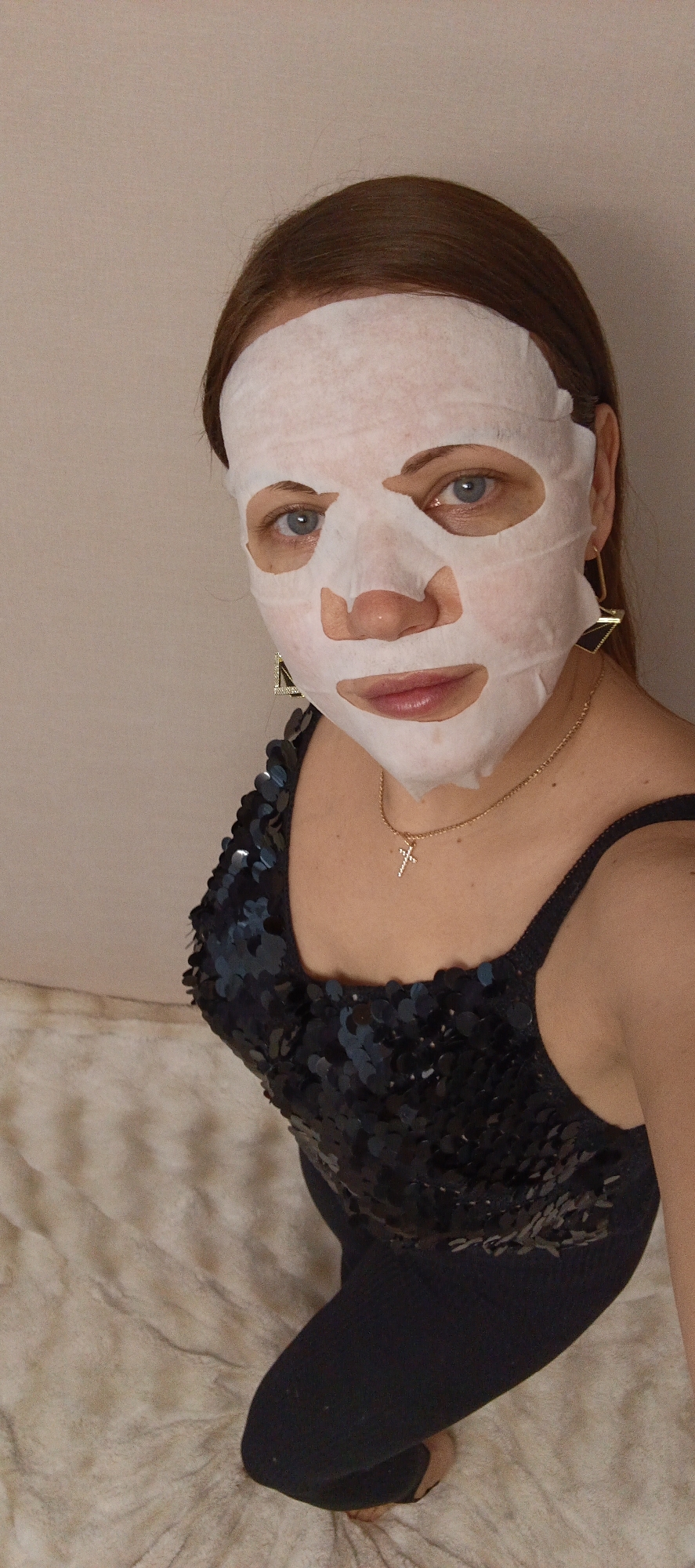 A close-up, eye-level shot captures a woman indoors, located in Chaikovsky, Russia, wearing a white sheet face mask. Her reddish-brown hair is pulled back, and her blue eyes are visible through the mask's cutouts. Her lips, also visible, are a soft pink color. She is wearing a dark-colored, possibly black or navy, sequined top or dress with spaghetti straps. A delicate gold chain necklace with a small, sparkling cross pendant adorns her neck. On her right ear, a distinctive large, geometric gold earring, shaped like a triangle with a dark insert, is visible.
The woman is looking directly at the camera with a neutral expression, suggesting she is posing for the photo while engaged in a self-care or beauty routine involving a facial mask. The background on the left consists of a plain, light beige or off-white wall. On the right, a soft, textured, light-colored surface, possibly a blanket or upholstered furniture, can be seen. The lighting in the image suggests an indoor setting with ambient illumination. There is no visible text in the image.