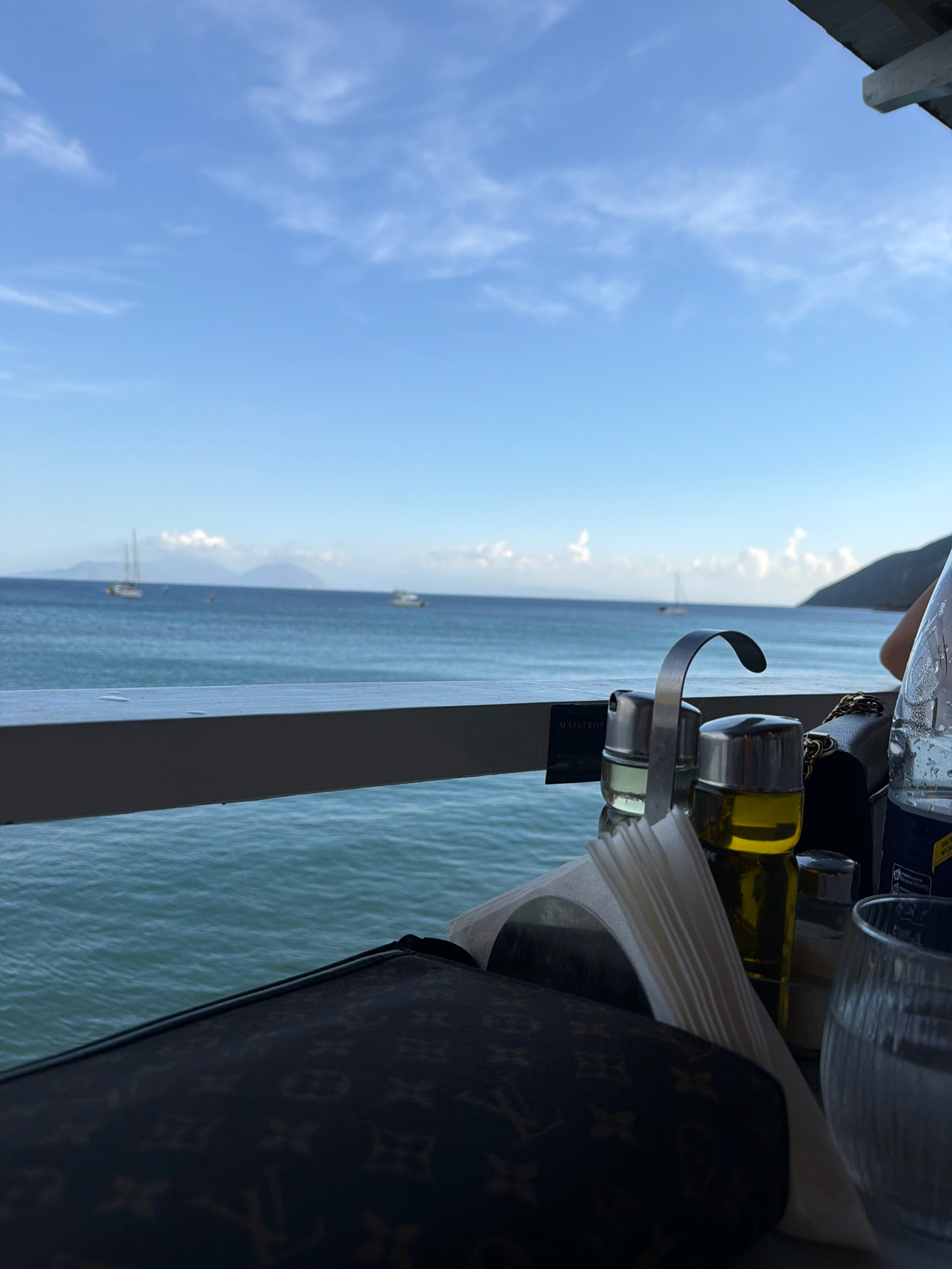The image depicts a table set for a meal at a restaurant overlooking the sea in the Municipal Unit of Apollonioi, Greece. On the table, in the foreground, are a patterned handbag, a napkin dispenser with white napkins, two glass bottles containing oil and possibly vinegar, a salt shaker, and a glass of water. A metal utensil holder is positioned behind the bottles. In the background, the turquoise sea stretches to the horizon, with several sailboats visible in the distance. Mountains form a backdrop against a clear blue sky dotted with white clouds. A railing separates the table area from the sea. The overall impression is one of a serene coastal dining experience.