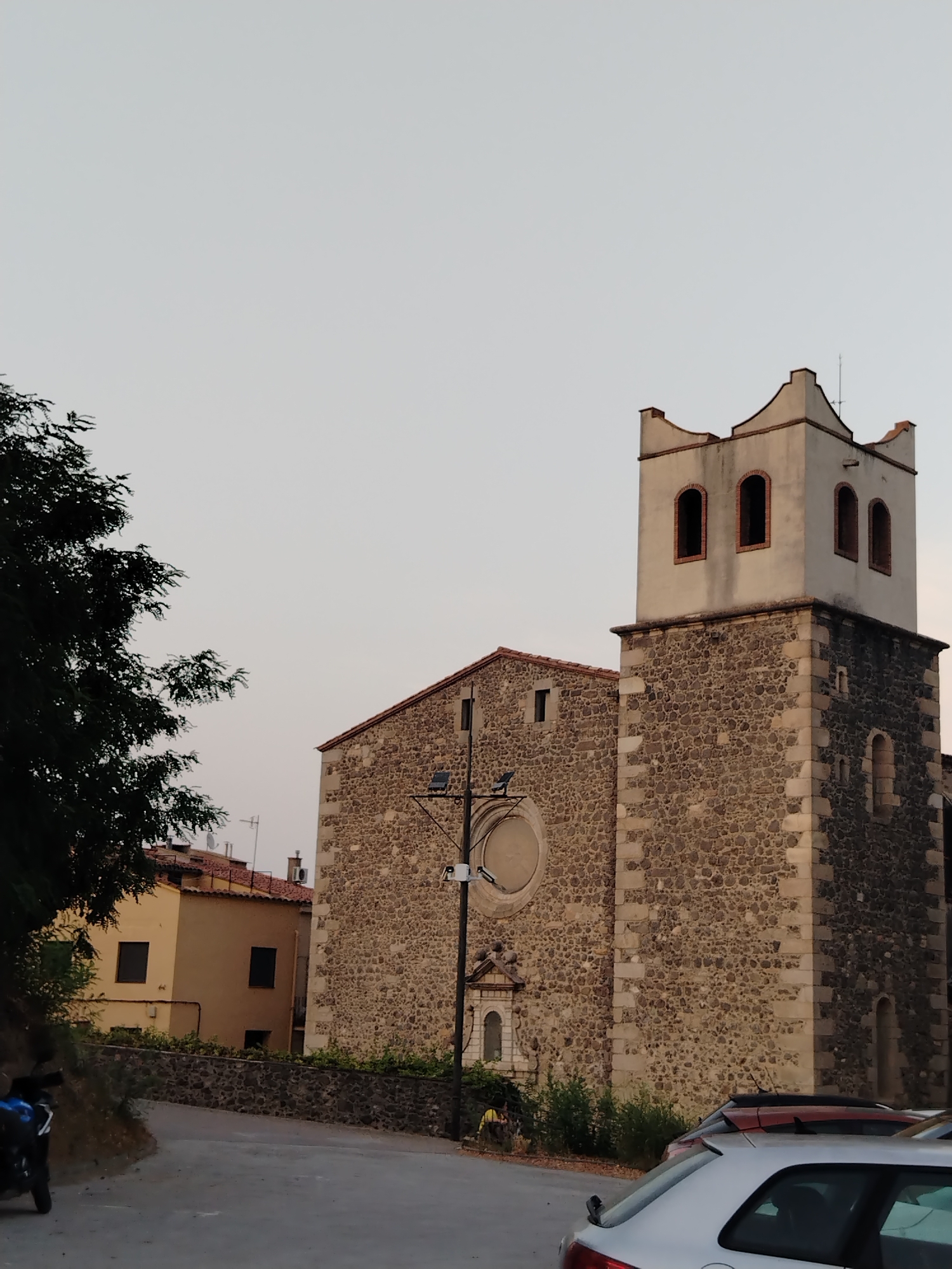 The image depicts the church of Santa Maria de les Arenes in Hostalric, Spain, viewed from a street. The church is constructed of stone with a prominent bell tower. The bell tower has a cream-colored top section with arched openings and a decorative crenellated edge. Adjacent to the church, a lighter-colored building with red-tiled roofs is visible. The street in the foreground is paved and curves to the left. A dark-colored tree with dense foliage is situated on the left side of the image, partially obscuring the sky. Several parked cars are visible on the right side of the street, in front of the church. The sky is overcast and a pale grey color. The overall scene suggests a quiet, daytime setting in a town.