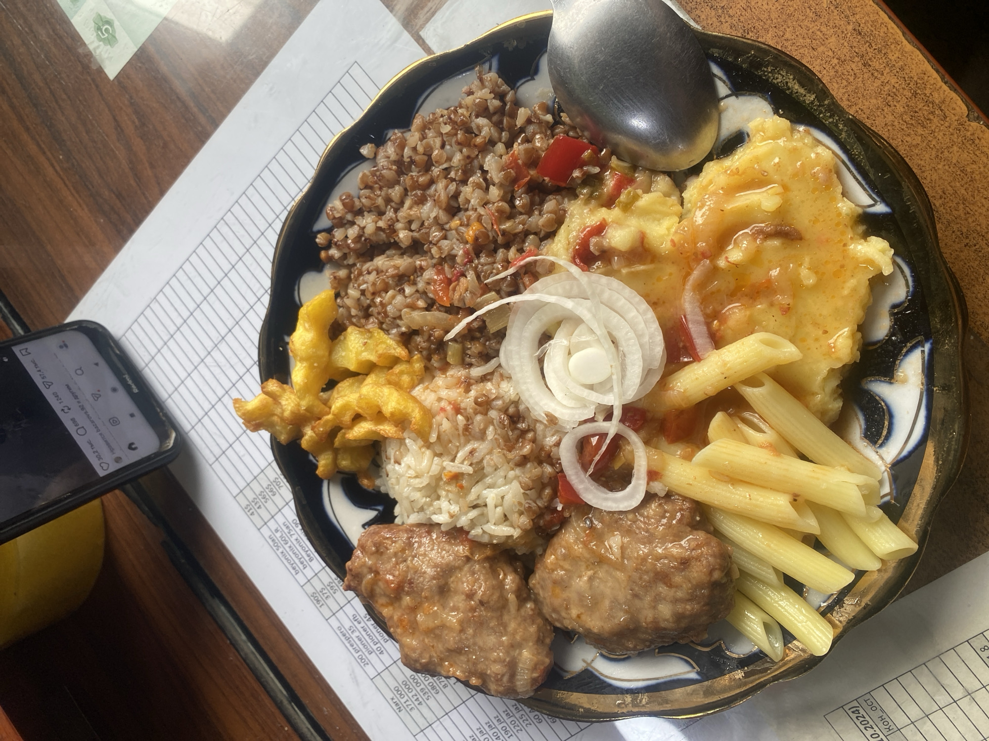 The image shows a plate of food, possibly a meal in Bukhara, Uzbekistan. The plate contains a variety of dishes including buckwheat, rice, fried potatoes, two meat patties, pasta, and a yellow, possibly egg-based dish. Sliced onions adorn the center of the plate. A spoon rests on top of the food. The plate itself has a blue and gold pattern. A smartphone lies on the left side of the image next to the plate, atop a printed sheet with rows and columns, likely a bill or an accounting document. The table has a wooden surface. The printed sheet has some text visible. The location is likely a restaurant or a café in Bukhara, Uzbekistan, where such dishes are commonly served.