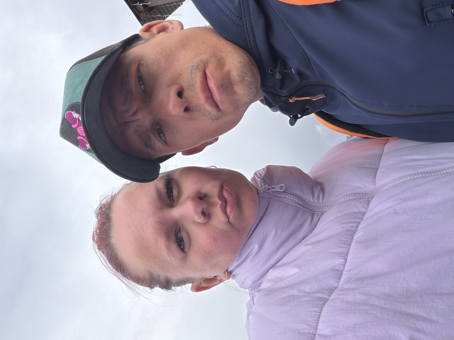The image features two individuals, a man and a woman, captured from an extreme low-angle perspective. The image is rotated 90 degrees clockwise, showing their faces close-up as if they are looking directly down at the camera. The man, positioned towards the top of the frame, wears a dark blue jacket with orange accents and a dark baseball cap with a green brim and an indistinct pink/purple graphic on the front. He has short facial hair and a neutral expression.
Below him, the woman wears a light purple puffer jacket. She has reddish hair and a silver nose ring on her left nostril, displaying a neutral to slightly serious expression. The background consists of a light, overcast sky, indicating a cloudy daytime setting outdoors. A dark structure, possibly a roof or building, is subtly visible in the very top right corner of the frame. This scene appears to be an outdoor selfie taken in Chaikovsky, Russia. No discernible text is visible on any objects or clothing.