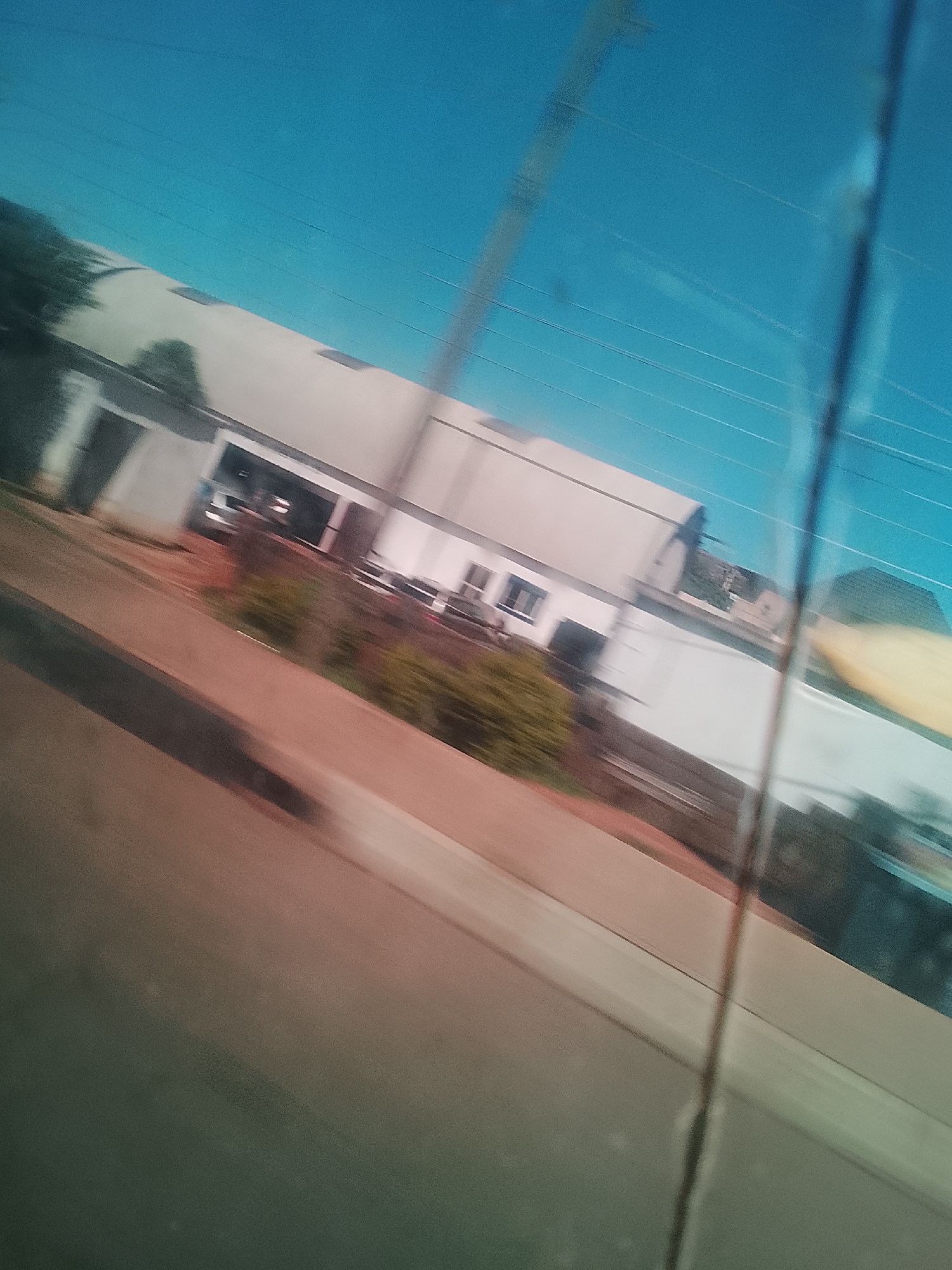 The image depicts a blurry, low-resolution, outdoor scene, likely taken from a moving vehicle. The sky is a bright, clear blue with thin, horizontal lines that may represent wires or power lines. Below the sky is a light-colored building with a curved roof, possibly an industrial or commercial structure. In front of the building is a patch of green foliage. A road or paved area runs diagonally across the lower portion of the image, with a reddish-brown hue. The overall impression is that of a fleeting glimpse of a city landscape. The exact location in Gura Top, Nigeria is not determinable from the visual information provided.