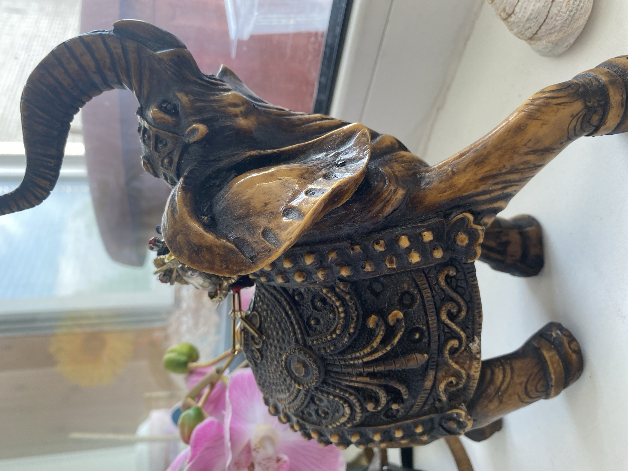 A close-up, eye-level, medium shot captures an ornate, brown, carved elephant figurine. The figurine is positioned on a white surface, possibly a windowsill, with a blurred background. To the left, a cluster of pink orchid flowers with green buds is visible. To the right, a portion of a seashell is present. The elephant figurine is richly decorated with intricate patterns on its saddle and body. Its trunk is raised, and its ears are spread. The lighting suggests an indoor environment with natural light coming from a window. The image does not contain any identifiable elements that would suggest its location in the city of Tyumen, Russia.