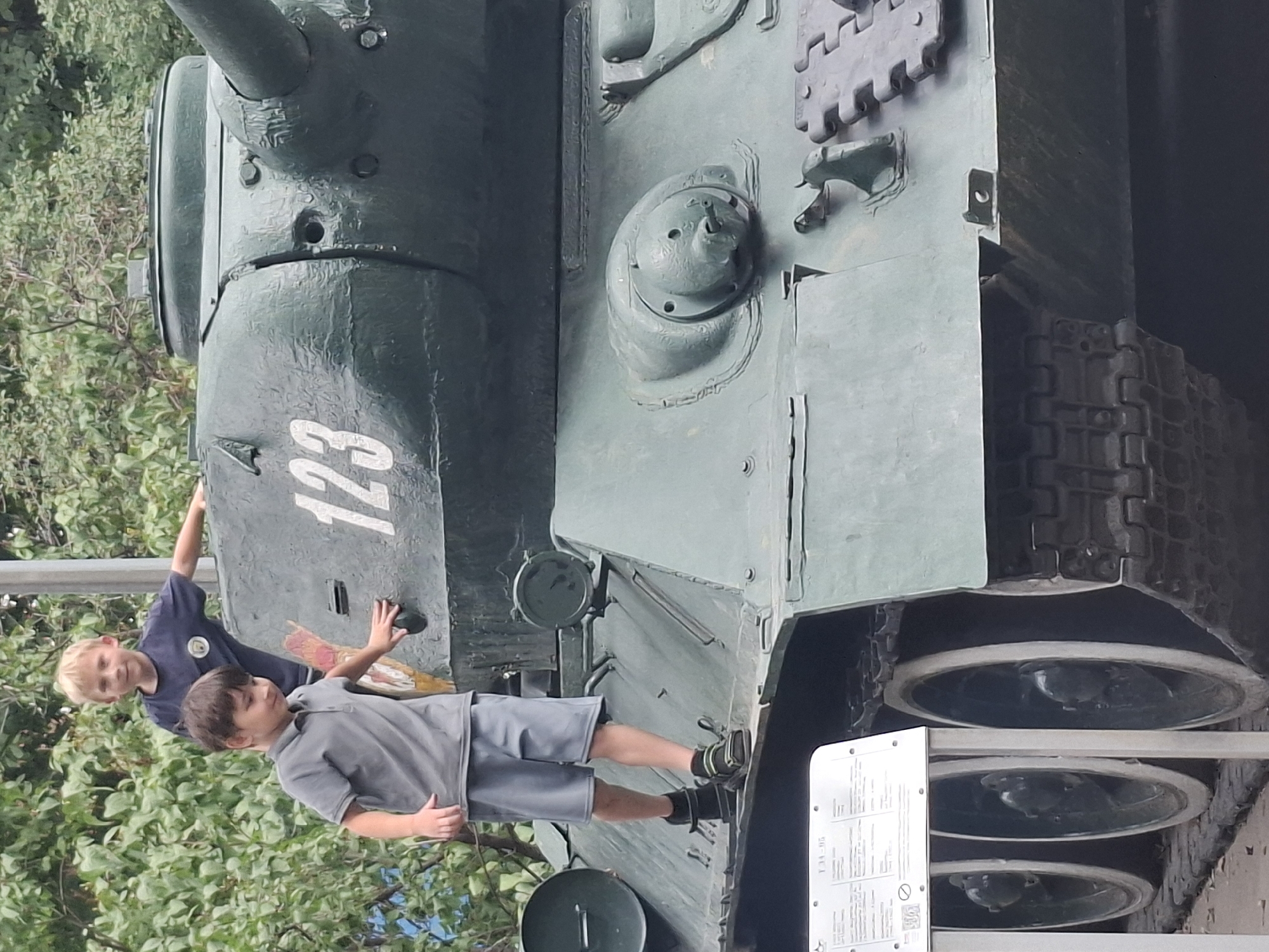 Two young boys stand on a green tank. The boy in the foreground, wearing a gray polo shirt and gray shorts, is on the tank's track. The boy in the background, wearing a dark blue t-shirt, is standing on the turret. The tank is positioned outdoors, with foliage and trees visible in the background. A sign with text and a logo is visible on the left side of the image. The tank is likely a T-34-85, given the markings on the sign. This image was taken at the Museum of Weapons and Military Equipment in Almaty, Kazakhstan.
