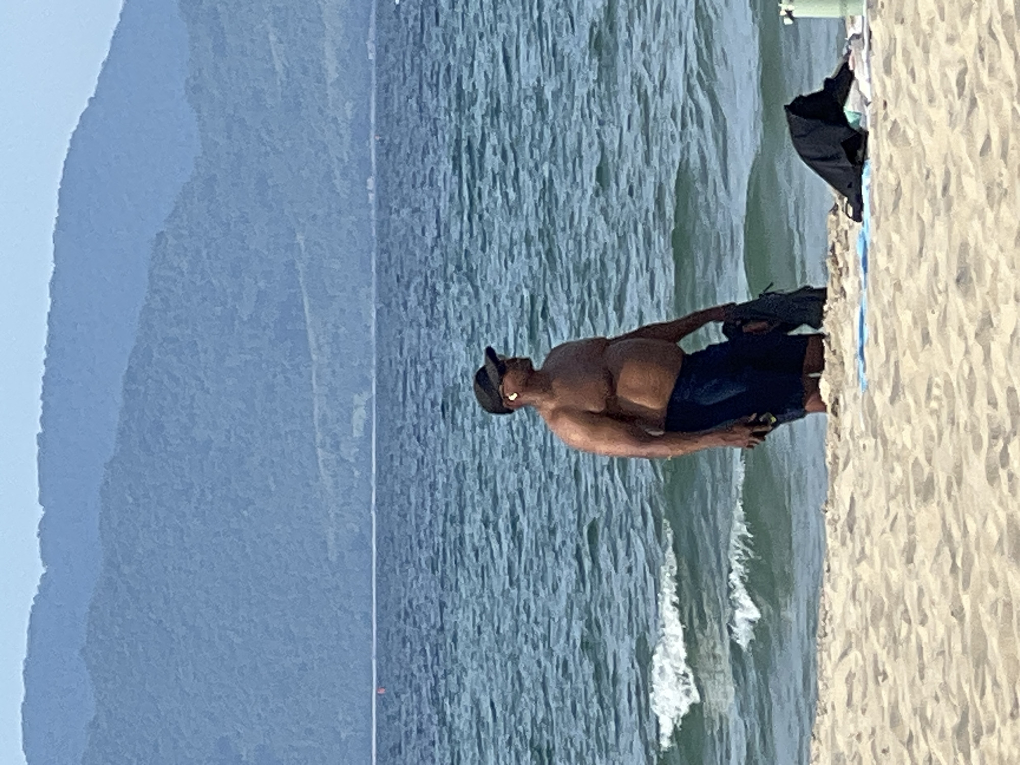 An adult male stands on a sandy beach with the ocean in the background. He is wearing dark blue shorts and a black baseball cap, and appears to have earbuds in. He is holding a dark colored garment in his left hand. The ocean has moderate waves and the sky is a hazy blue. In the distance, mountains are visible. A few items of clothing are visible on the sand near the man. The location is the coastline of Đà Nẵng, Vietnam.