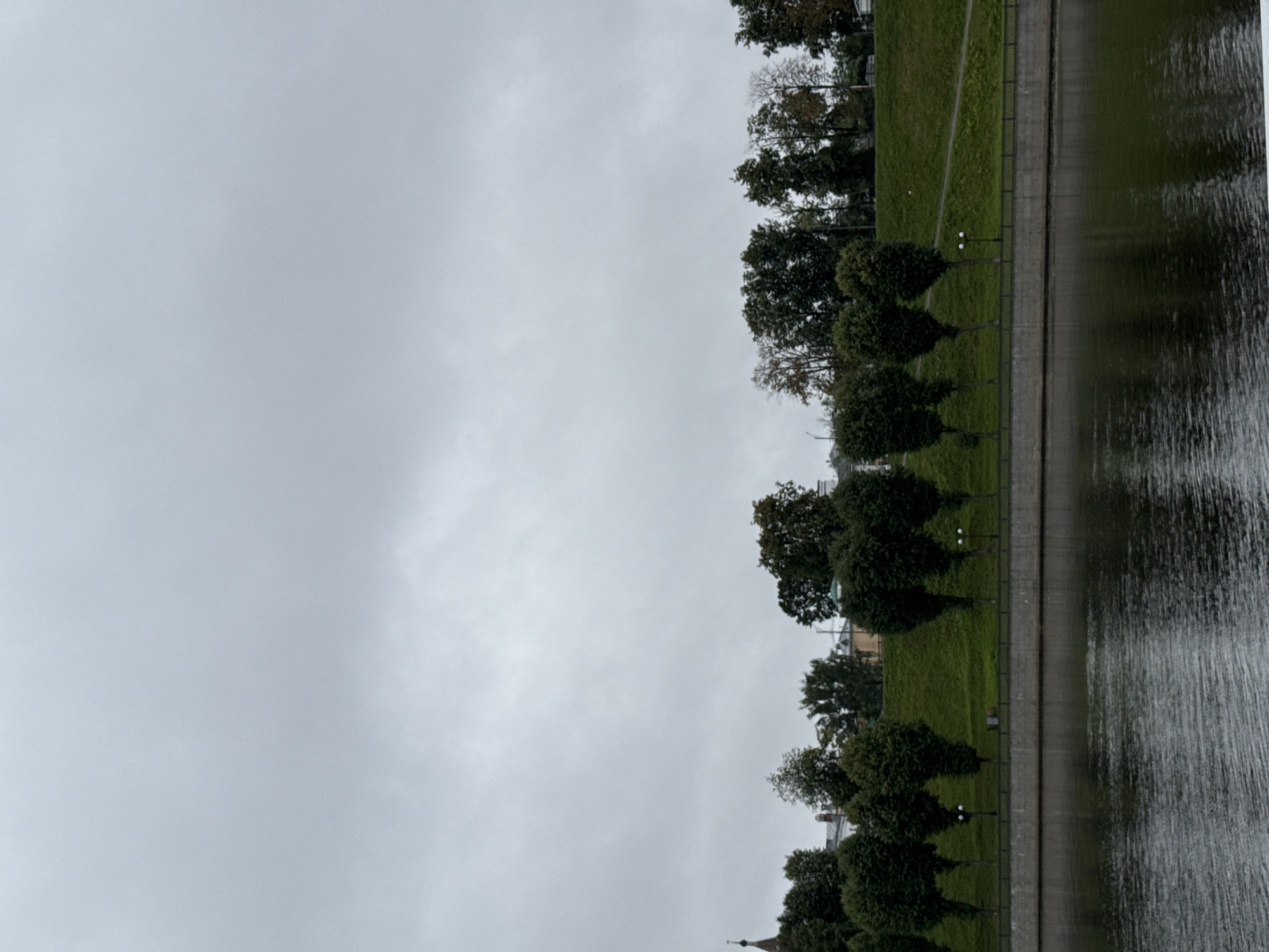 The image depicts a serene waterfront scene under a cloudy sky. In the foreground, a dark, rippling body of water reflects the overcast sky and the trees lining the opposite bank. A paved embankment separates the water from a gently sloping grassy hill.
Along the crest of the hill, a row of evenly spaced, manicured trees stands in stark contrast to the natural growth further up the slope. Beyond the trees, glimpses of buildings, including what appears to be a church spire, can be seen. The overall atmosphere is one of quietude and natural beauty.
The location is identified as Шебунино, Russia.