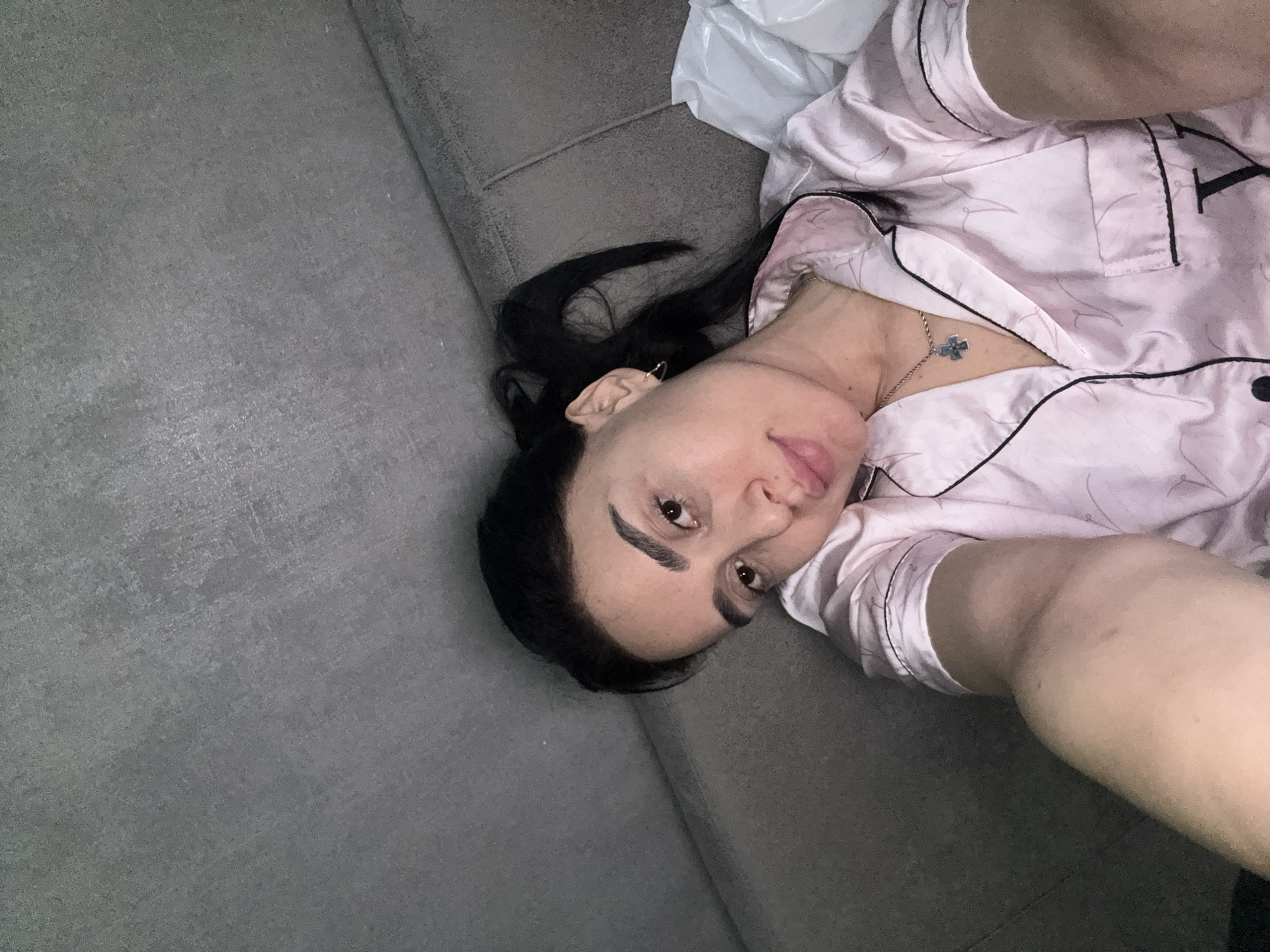 A woman is lying on a gray couch indoors. She has dark hair and fair skin, and she is wearing a pink pajama top with black piping. A silver necklace with a cross pendant is visible around her neck. A white plastic bag is partially visible near her head. The location is not determinable from the image.