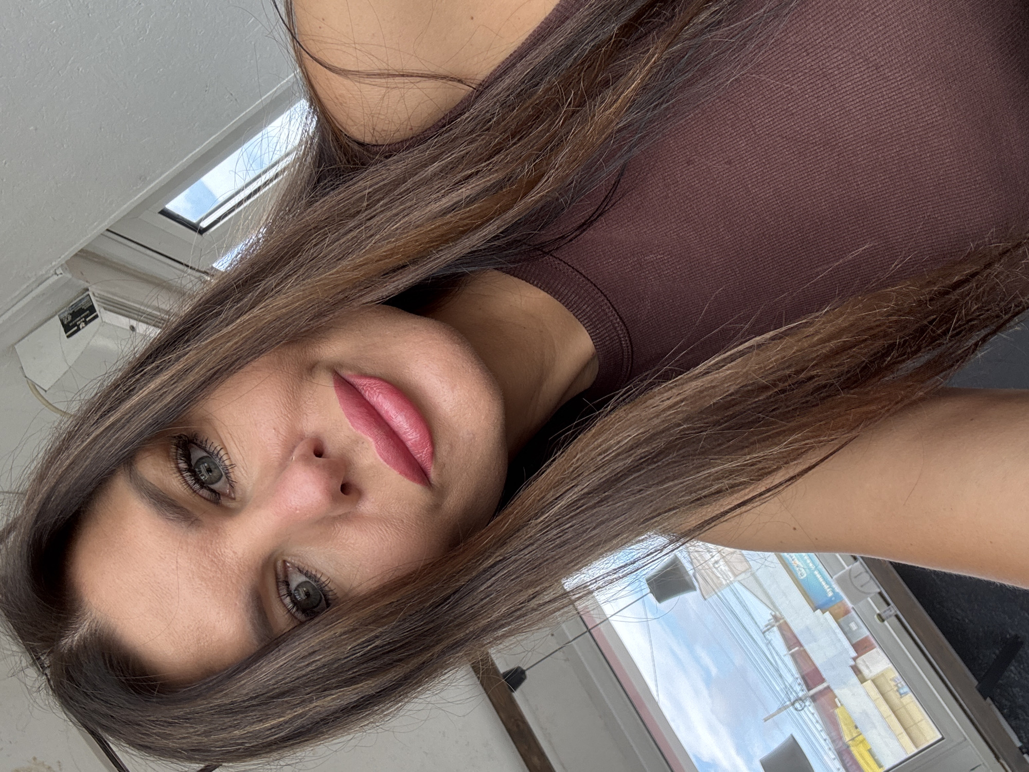 A close-up selfie of a woman with long, dark brown hair, green eyes, and full lips wearing pink lipstick. She is indoors, with a window in the background showing a cloudy sky and buildings. The woman is wearing a brown, ribbed tank top. The scene appears to be in the city of Chaikovsky, Russia, suggested by Cyrillic text visible in the background of the window.