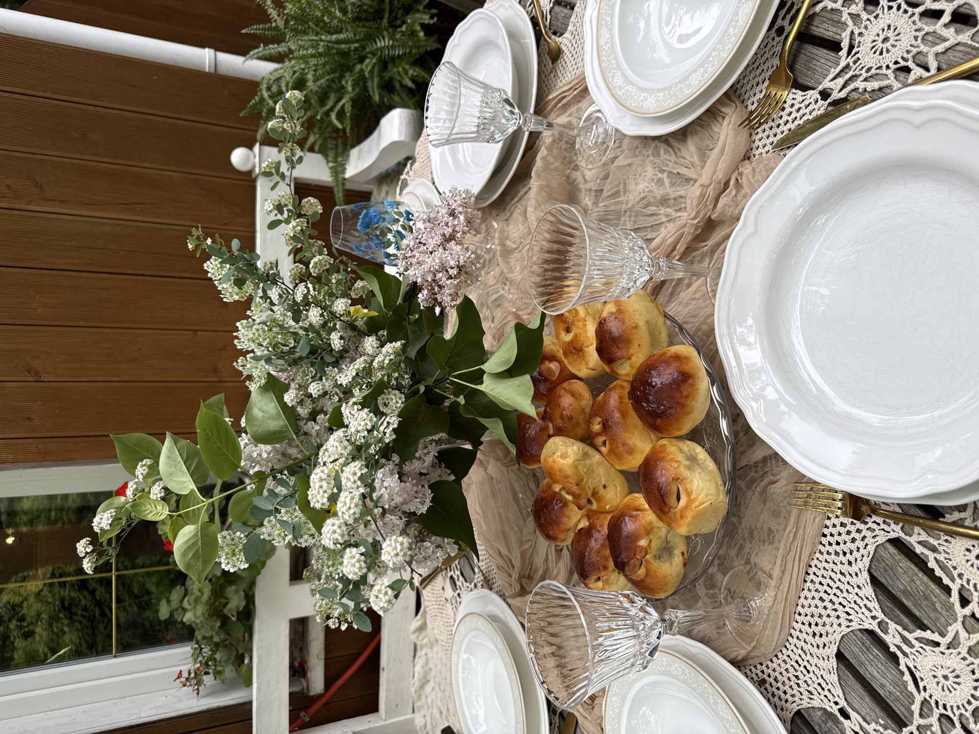 A detailed, top-down view captures an outdoor table setting, likely for a small gathering. The table is set with white plates, golden cutlery, and crystal glasses, arranged on a beige cloth draped over a wooden surface. A glass plate filled with baked goods rests near the center of the table. A floral arrangement in a blue patterned vase provides a natural, decorative touch. The location appears to be a veranda or outdoor seating area, adjacent to a wooden-clad wall with a window. The scene is illuminated by natural light. The location is inferred to be Sapronovo, Russia. There is no indication of any people in this scene.