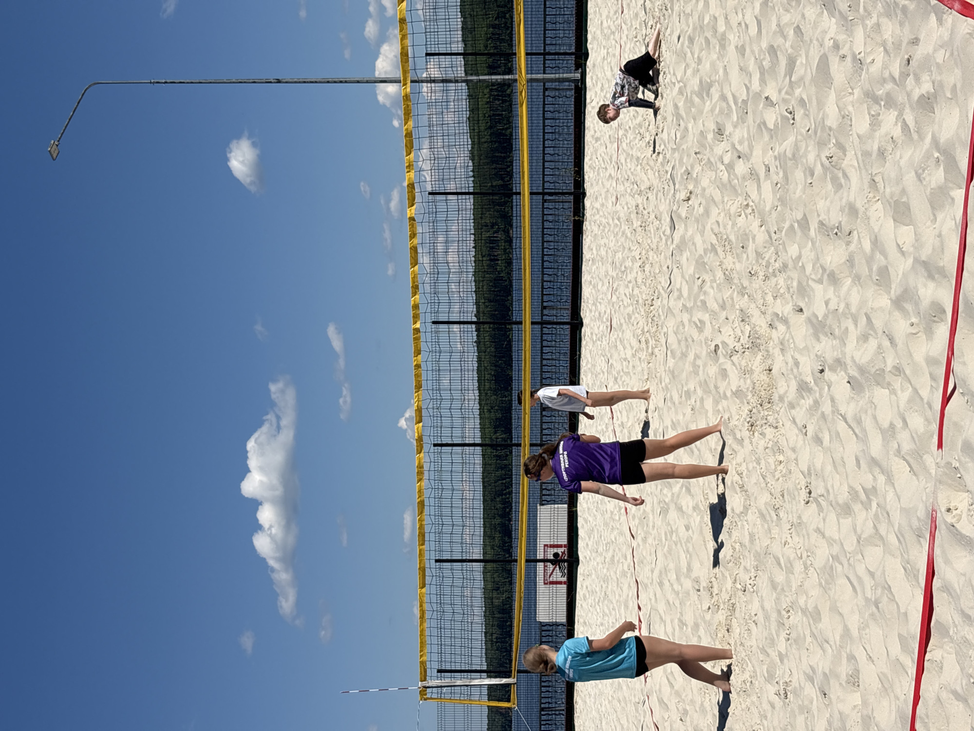 This is a wide-angle, eye-level shot of a beach volleyball game on a sunny day at the Kama River embankment in Chaikovsky, Russia. Four young people are visible on the sand court. On the left, a girl in a turquoise t-shirt and black shorts stands, facing away from the camera. To her right, a girl in a purple t-shirt and black shorts also stands, facing away from the camera. Further to the right, a person in a white t-shirt and black shorts is standing. In the background, on the far right of the court, a person in a patterned t-shirt and black shorts is crawling on the sand, looking towards the net. The volleyball net, with yellow top and bottom tape, is stretched across the court. A red boundary line is visible on the sand in the foreground. Behind the court is a dark fence, and beyond that, a wide body of water, identified as the Kama River, with a tree-covered shoreline in the distance. The sky is a bright blue with scattered white clouds. A tall lamp post is visible on the right side of the frame.