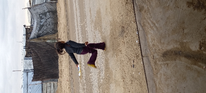 A young child, appearing female with dark hair, is captured mid-stride running from right to left across a dusty, unpaved road in Khan Yunis, Palestinian Territory. The child wears a dark long-sleeved top, dark purple pants, and light-colored shoes, holding a small, multi-colored object in her left hand. The foreground consists of dirt and scattered debris, with a dark, wet-looking patch visible at the bottom right. In the background, several makeshift structures, appearing to be shelters or tents, are constructed from corrugated metal, wood, and various tarpaulins or blankets, one featuring a dark blanket with a light floral pattern. The sky is overcast and light grey.