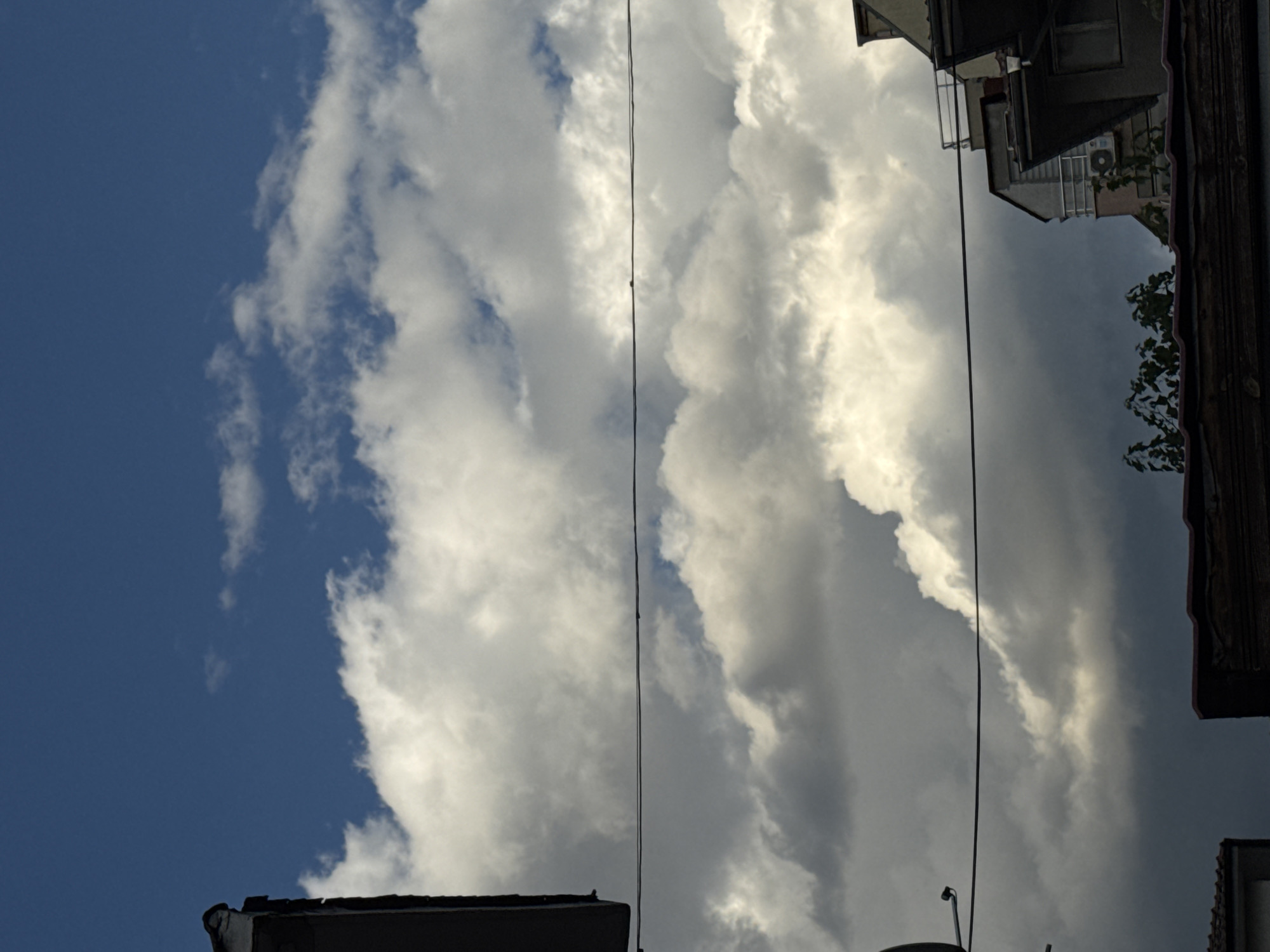 The image shows a view of the sky with clouds over buildings in Plovdiv, Bulgaria. The sky is a deep blue with white and gray clouds. There are power lines stretched across the frame. The buildings are partially visible at the bottom and edges of the image, with rooftops and parts of their facades showing. The architecture appears to be a mix of older and more modern structures. Some greenery is visible in the lower portion of the image. The overall impression is of an urban landscape viewed from below, with the sky and clouds dominating the scene.