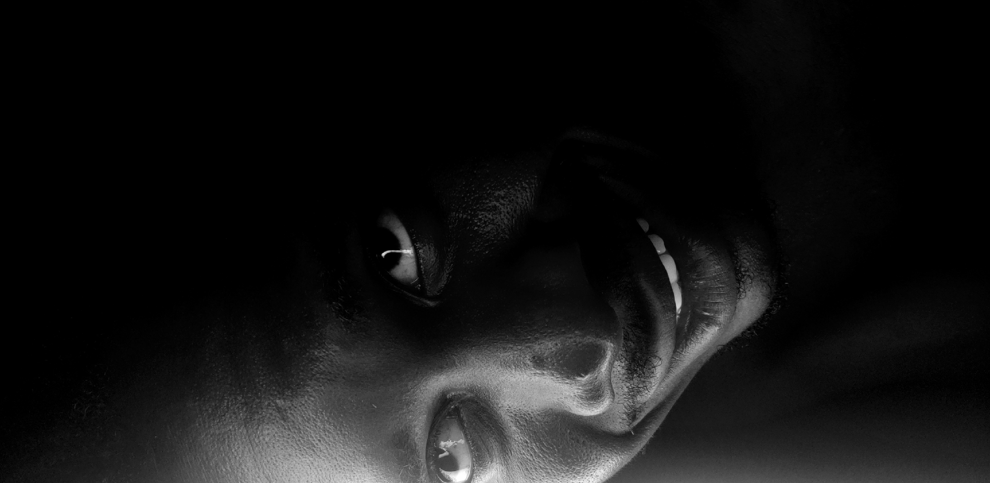 This media file is a high-contrast, monochromatic (black and white) close-up portrait of a person's face, set against an almost entirely dark background. The image is taken in Narayi, Nigeria.
The subject is an individual, likely an adult, whose facial features emerge from deep shadows. The composition shows the face from an unconventional angle, possibly looking upwards or with the head tilted back. The top and left portions of the frame are consumed by blackness, with details becoming visible towards the bottom and right.
Notable details include the person's eyes, which are open. The left eye (viewer's left) prominently displays the white of the eye and a distinct highlight, indicating a strong, focused light source. The right eye is also visible, though less illuminated. The mouth is slightly agape, revealing a row of upper teeth that contrast sharply with the dark skin, suggesting an expression that could be a subtle smile, a gasp, or contemplation. Fine textures of the skin are discernible where the light catches the surface, and some short, dark hair or stubble is visible along the jawline/cheek.
No specific activity or event is clearly depicted beyond the portraiture itself. The extreme darkness of the scene makes it impossible to determine the time of day or any outdoor weather conditions. There are no visible objects or discernible text within the image. The overall mood conveyed by the dramatic lighting and deep shadows is intense, intimate, and enigmatic.