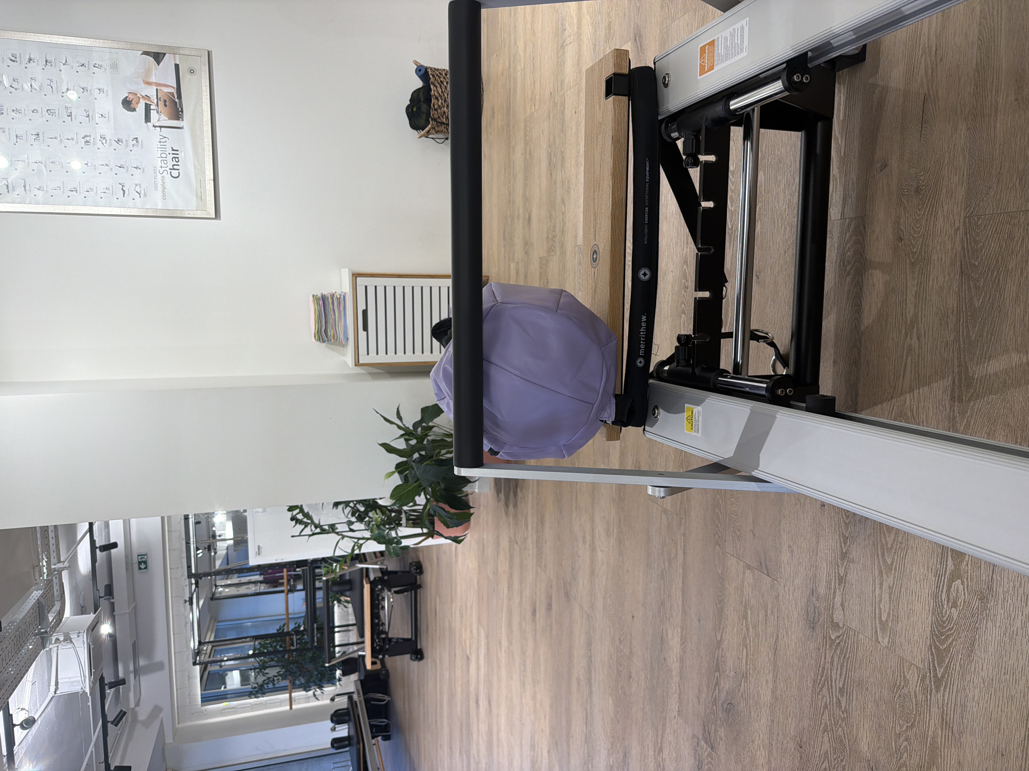 The image depicts an indoor space, likely a fitness studio or gymnasium, with a focus on Pilates equipment. In the foreground, a piece of Pilates apparatus is partially visible, featuring a metallic frame, a wooden platform, and a light purple upholstered component. A black horizontal bar runs across the top of the apparatus. The flooring is a light brown wood laminate. In the midground, there are several other Pilates reformers and chairs arranged in the room. Plants are visible in pots, adding a touch of greenery. The background shows a wall with a framed poster displaying exercise diagrams and text that reads "STOTT PILATES complete Stability Chair". A white cabinet with slatted doors is also present, topped with a stack of folded towels. Further back, through an opening, another part of the studio is visible with more equipment and mirrors. The lighting appears to be artificial, with ceiling lights and track lighting visible. There are no people visible in the image. The location is not Boreti, Montenegro, as there is no visual information to support this.