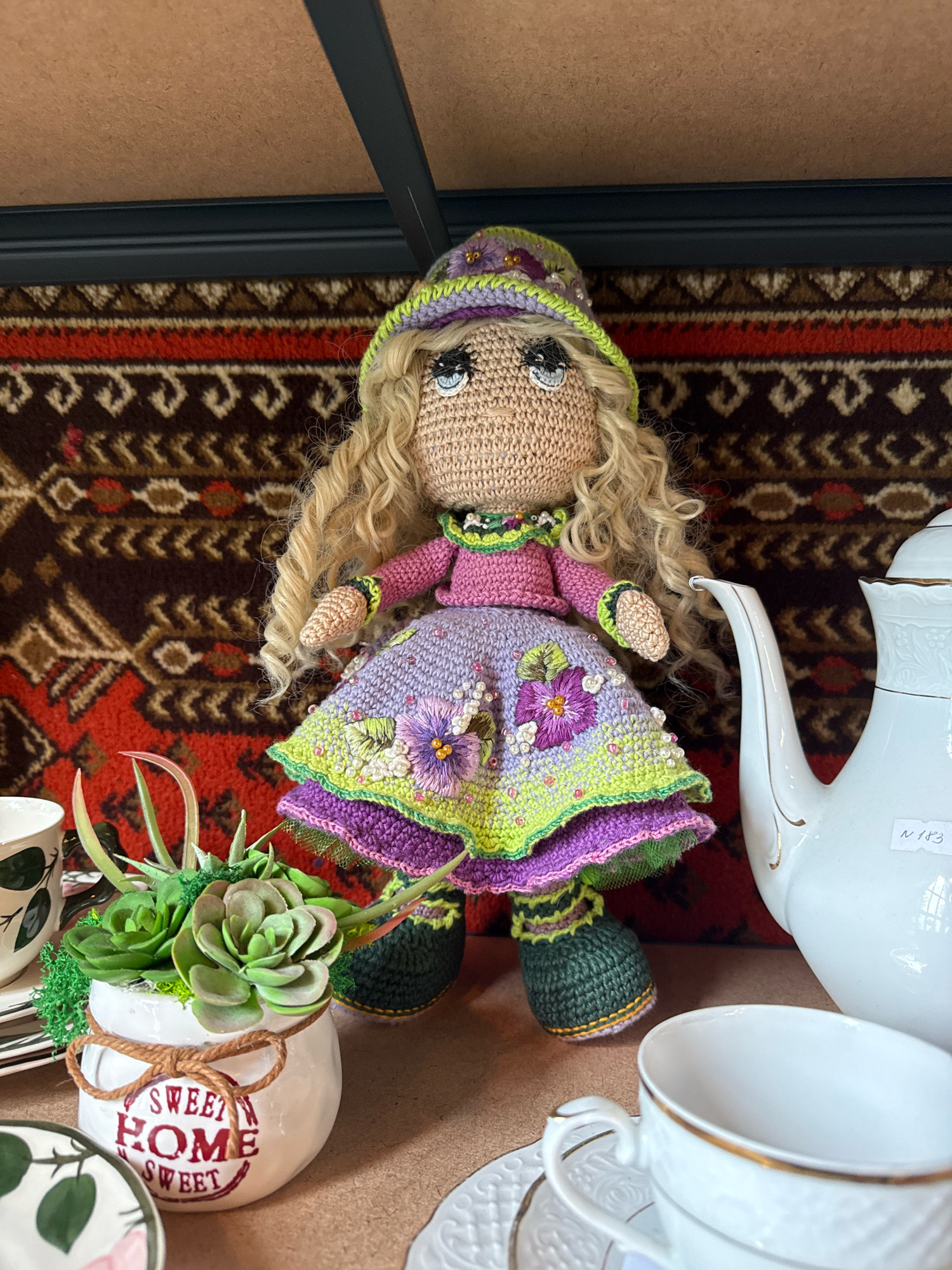 An indoor still life composition features a crocheted doll standing centrally on a surface, possibly a shelf or table. The doll is dressed in a purple and green outfit with a hat, and has blonde, curly hair. To the right of the doll is a white teapot with gold trim, and to the left, a white ceramic pot containing succulents. Several pieces of white teaware, including cups and saucers, are also visible. The background consists of a dark, patterned fabric, possibly a rug or tapestry. The scene appears to be domestic. There is no indication of Svetlogorsk, Russia.