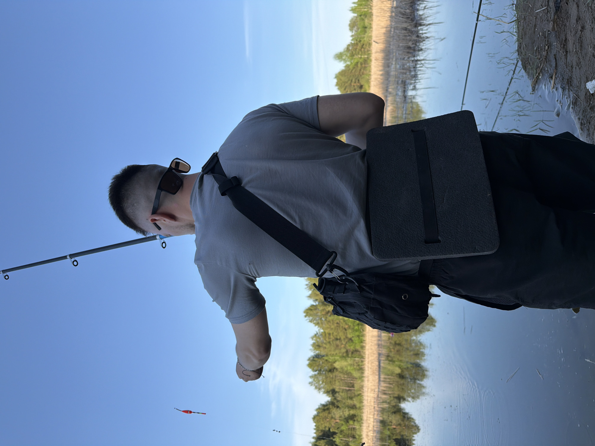 A person wearing a grey t-shirt, black sunglasses, and a black bag over their shoulder is fishing. They are standing next to a body of water with trees in the background. A fishing rod is extended with a red and white bobber in the air. The location is Unknown, Unknown.