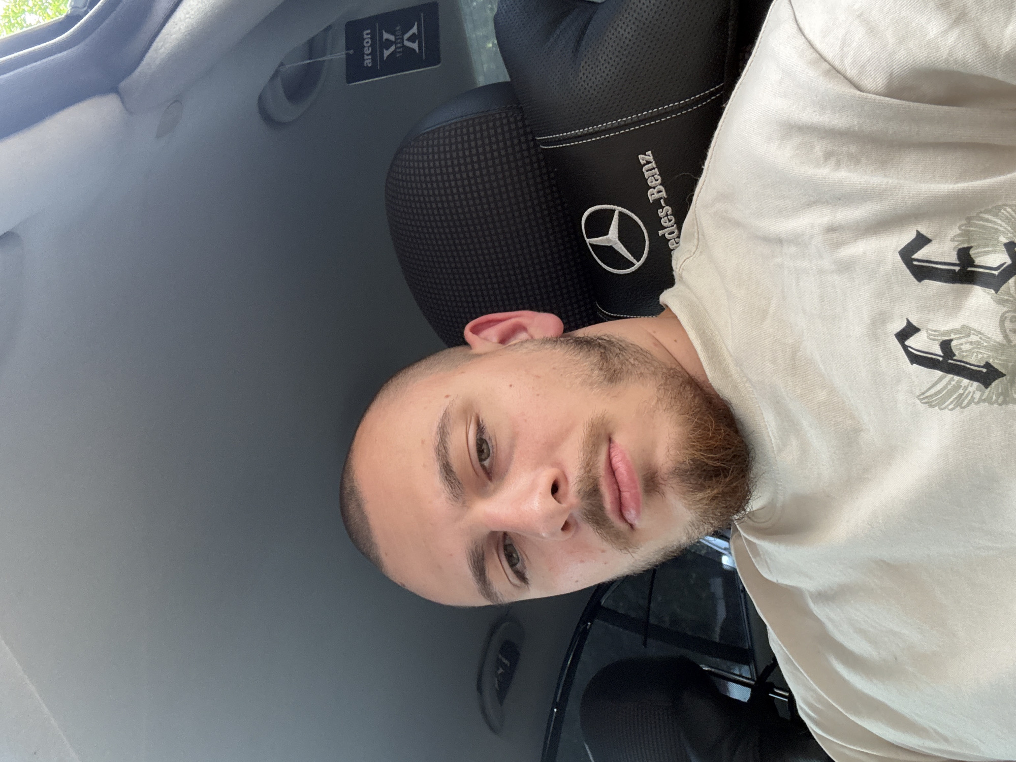 A selfie photograph taken inside a vehicle. The subject is a young male with a shaved head and a short beard. He is wearing a light-colored t-shirt. Behind him is a car seat with a Mercedes-Benz logo on the headrest. An air freshener is hanging from the rearview mirror. The environment appears to be the interior of a car. The photograph was taken in Stara Zagora, Bulgaria.