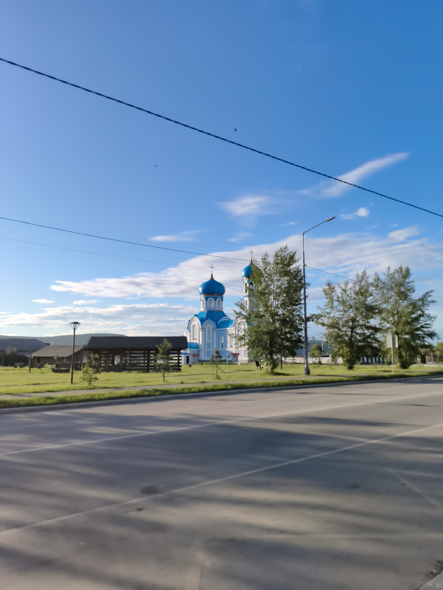 The image captures a sunny day in Svirsk, Russia, with a bright blue sky dotted with scattered white clouds. In the foreground, a paved road with white lane markings stretches across the frame, casting long shadows from unseen trees. To the left of the road, a neatly trimmed green lawn borders a sidewalk. Behind the lawn stands a light pole. Further back, a white and blue Orthodox church with a prominent blue dome and cross is visible. Adjacent to the church, several trees with lush green foliage are present, with a streetlamp situated between them. A wooden structure, possibly a pavilion or shelter, is also visible on the grassy area. In the background, a few low-lying buildings and a distant treeline can be seen under the vast sky. There are no people visible in the image.