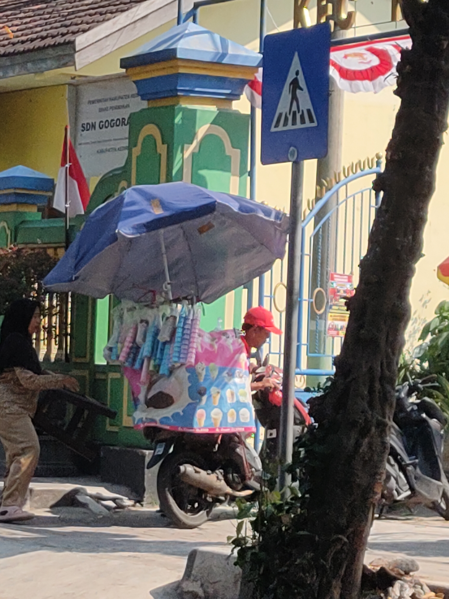 The image depicts a street scene in Ngasem, Indonesia, on a sunny day. In the foreground, a motorcycle with an attached cargo box is parked, adorned with a colorful print featuring ice cream cones and cartoon cows. A blue umbrella shades the cargo area, from which hang numerous small items, possibly snacks or toys. A person wearing a red cap and a red shirt is tending to the motorcycle. To the left, a woman wearing a black hijab and a patterned brown and white outfit is standing near a set of stairs. Behind them, a building with a yellow facade and green accents is visible, with a sign that reads "SDN GOGORAN." A pedestrian crossing sign is present on the right side of the image, and a large tree trunk with textured bark is situated in the right foreground. In the background, a blue metal gate and lush green foliage are apparent. Indonesian flags, red and white, are also visible.